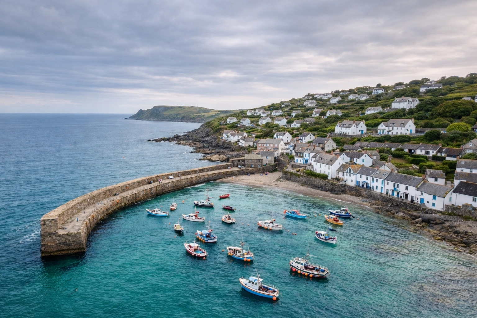 A high-resolution, scenic aerial photograph of Coverack village in Cornwall, UK. The image shows the iconic stone harbour wall, colourful fishing boats bobbing in the clear turquoise water, and traditional whitewashed cottages along the rugged coastline.