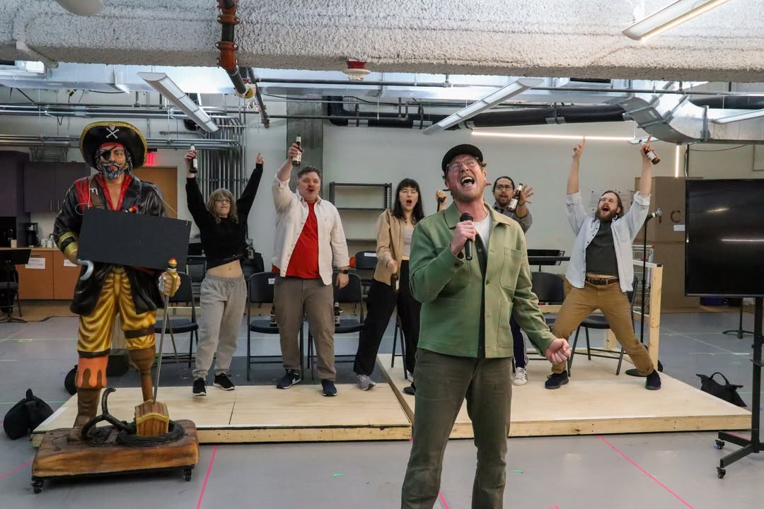 A group of artists rehearses a new performance in a studio One person sings passionately at the front while others act energetically behind on a platform. A colorful pirate statue is on display at stage left, suggesting playful experimentation and interdisciplinary storytelling within The Orchard Project's creative incubator.