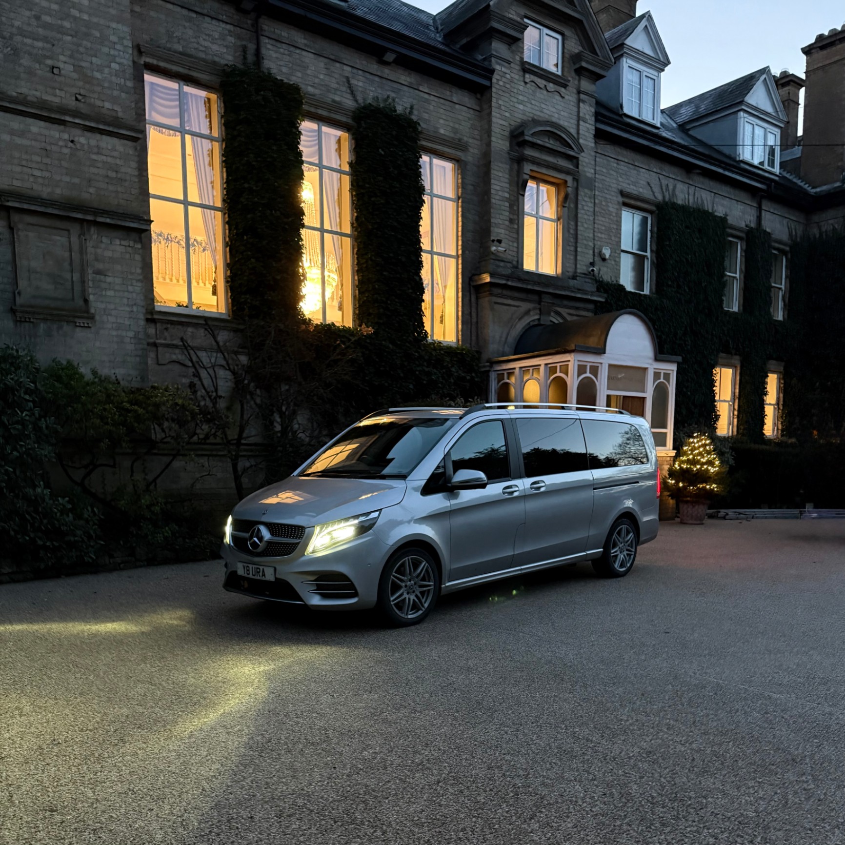 A Mercedes-Benz executive van from Aura Journeys is parked outside an elegant, illuminated manor, prepared for luxury chauffeur service.