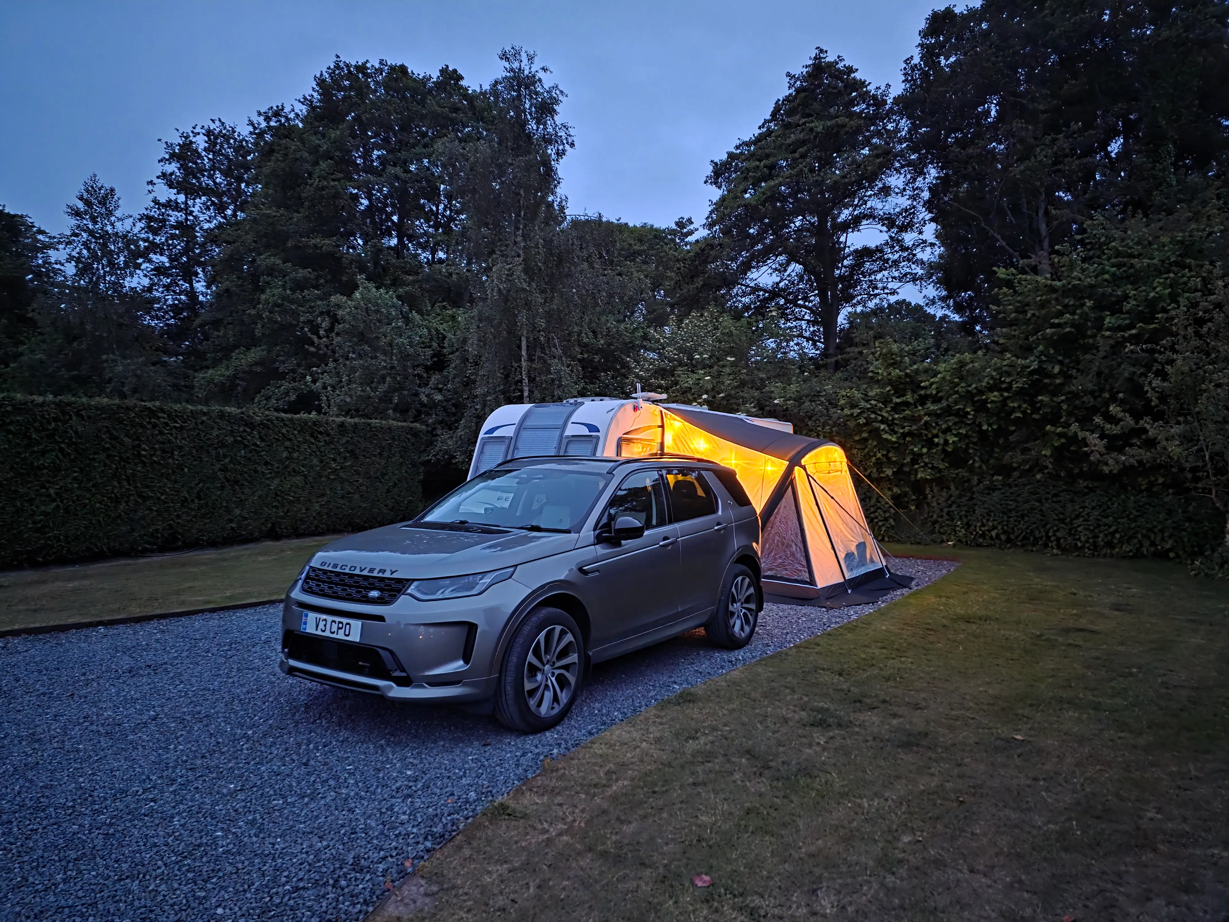 Evening setup at a gravel pitch featuring our Coachman Acadia 565 caravan with a fully erected awning, warmly lit from inside, and our Land Rover Discovery parked alongside. Surrounded by hedges and trees, this image showcases the peaceful atmosphere of a typical UK campsite perfect for caravanning reviews.