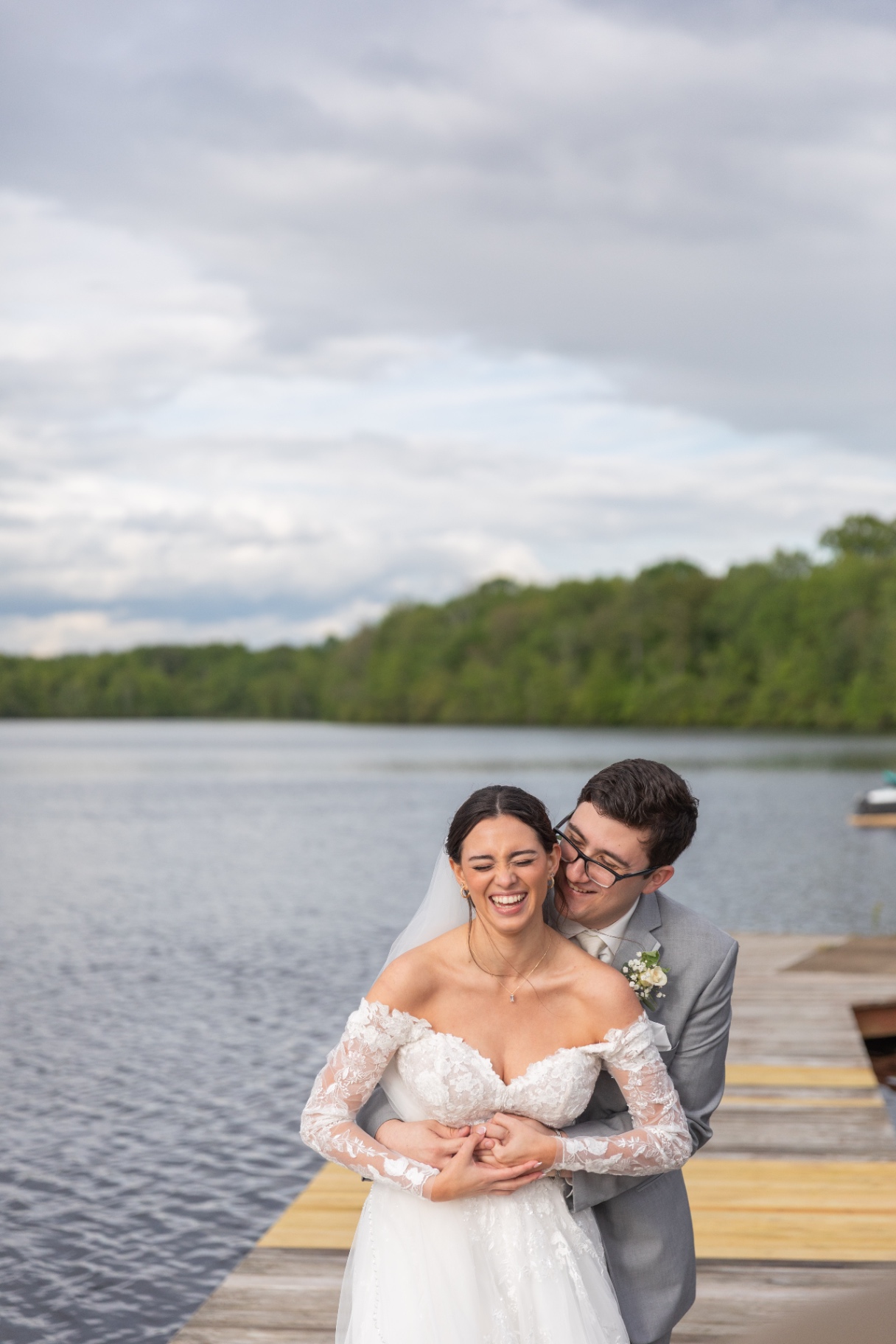 A bride and groom share a candid embrace and laughter on a wooden dock by the lake