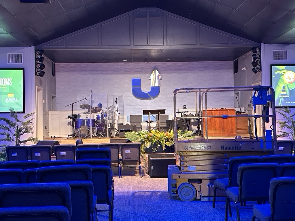 Sanctuary stage featuring a drum kit, lectern, and plants at the front. Newly installed SM Lights high-output LED fixtures provide front wash and even lighting coverage.