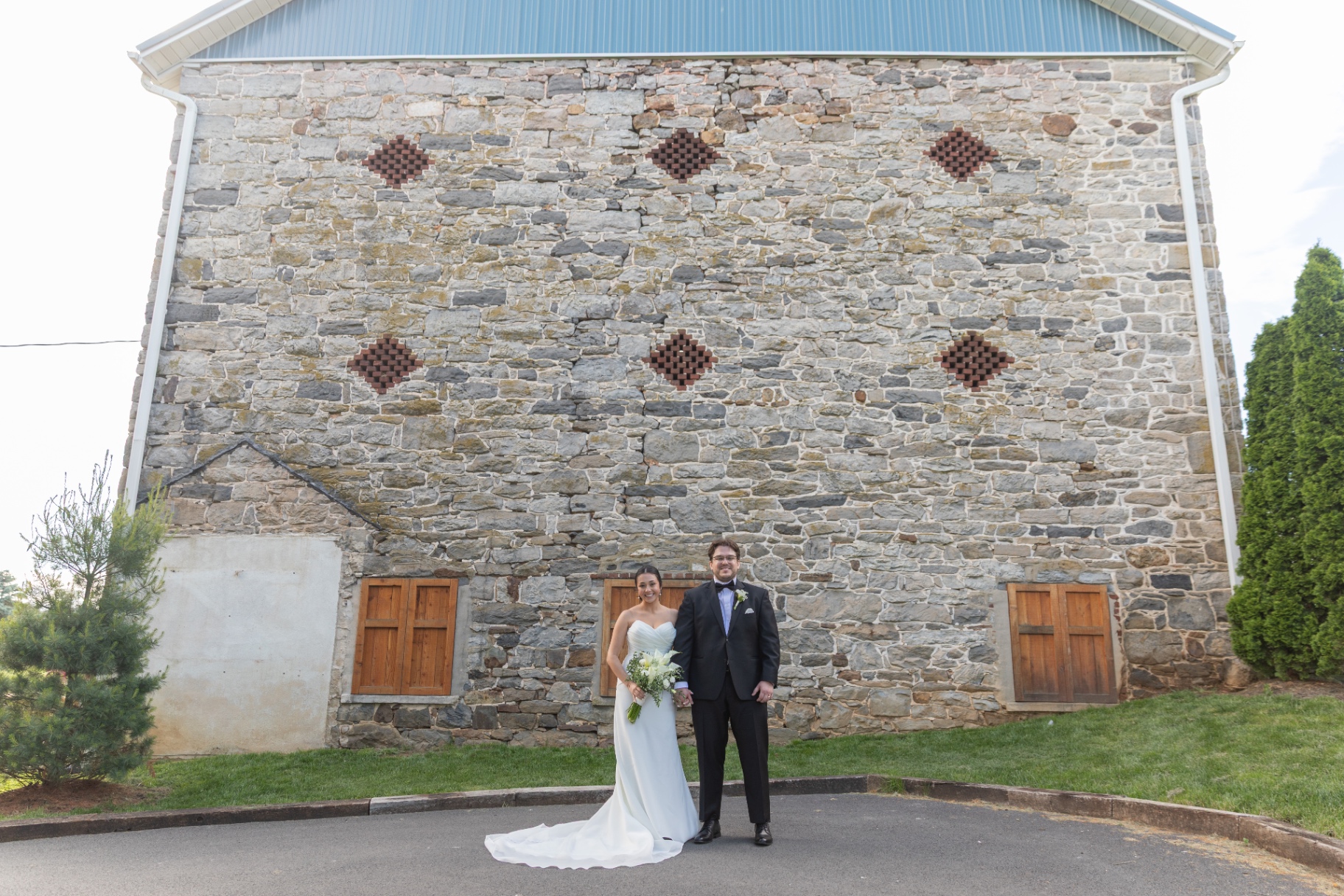 Bride and groom stand hand in hand in front of a rustic stone barn wall
