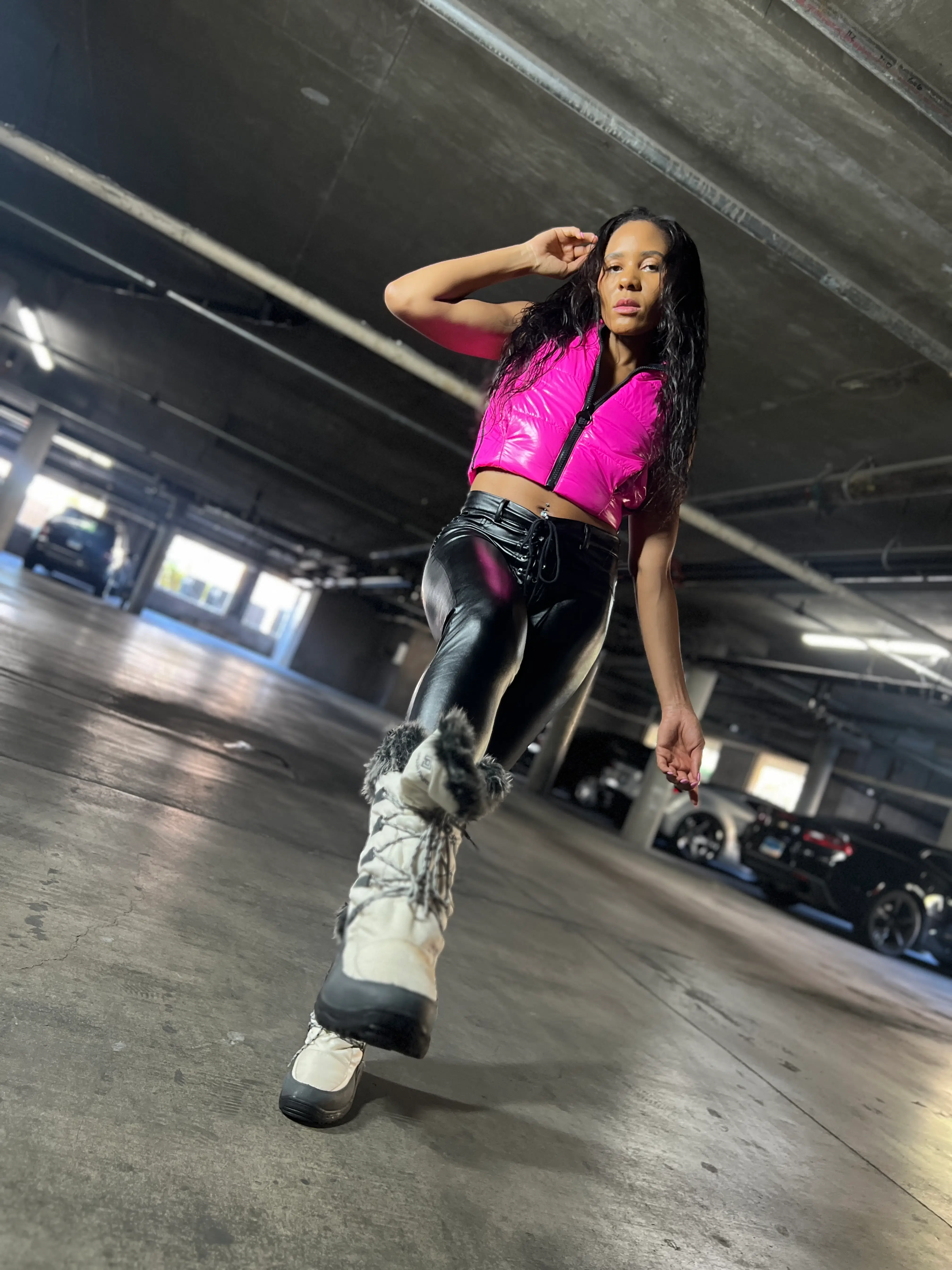 Model walks confidently in an urban parking garage, wearing a hot pink, cropped puffer vest and black faux leather leggings.
