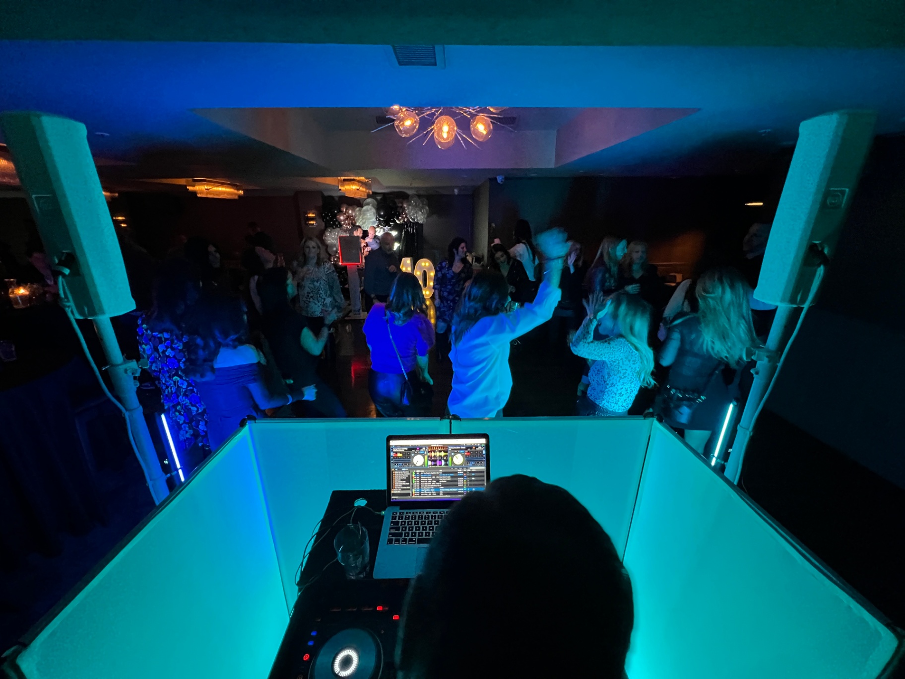 View from behind the DJ booth at Modish Entertainment event View from behind the DJ booth showing a lively dance floor with vibrant blue lighting, professional DJ equipment, and high-end speakers. Guests are dancing and celebrating amid well-coordinated decor, including illuminated balloon arrangements and marquee numbers, demonstrating Modish Entertainment's expertise in full-service event production and atmosphere creation.