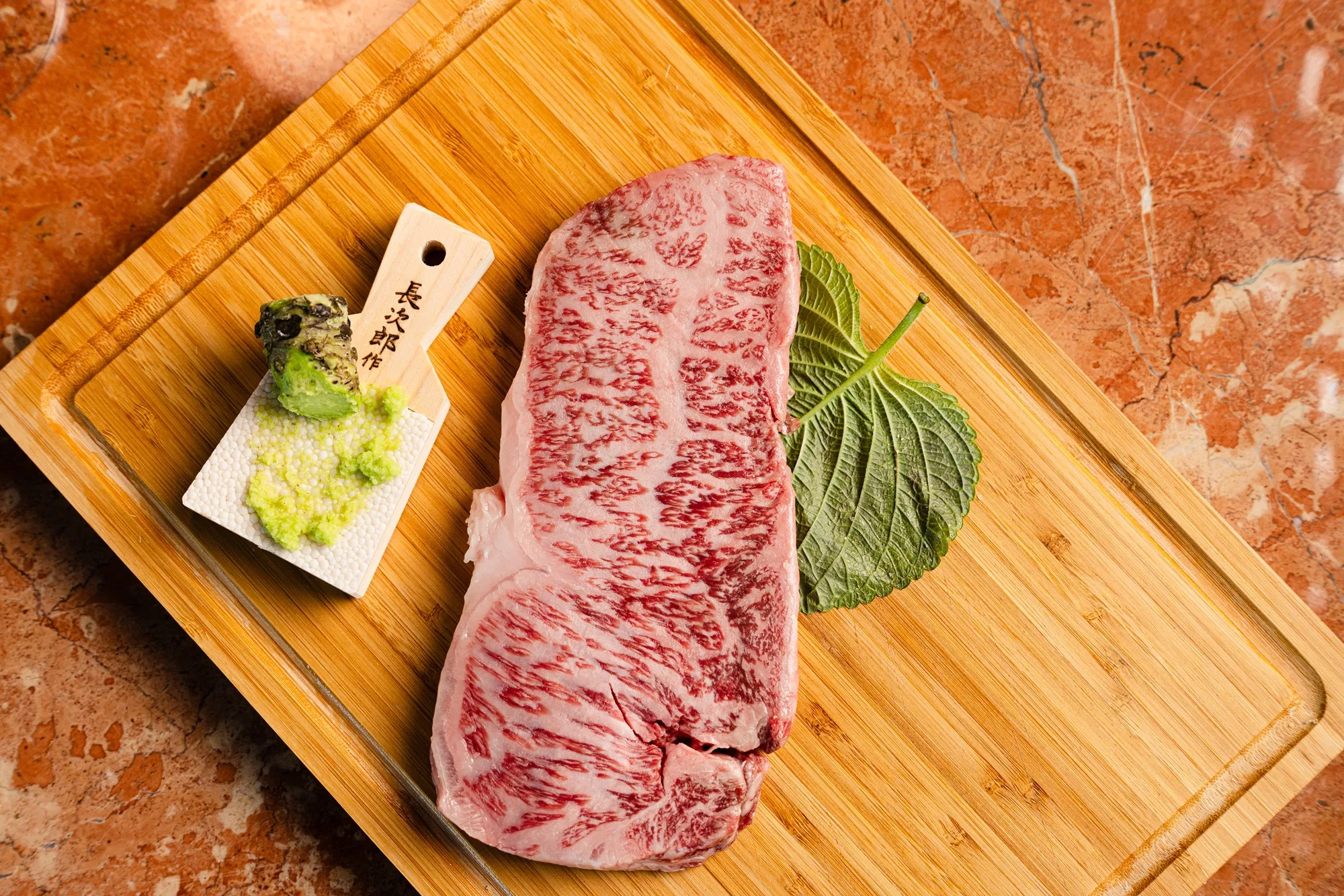 A beautifully marbled A5 Japanese Wagyu striploin steak sourced from Meat N' Bone, presented raw on a wooden board with a fresh shiso leaf and authentic wasabi root