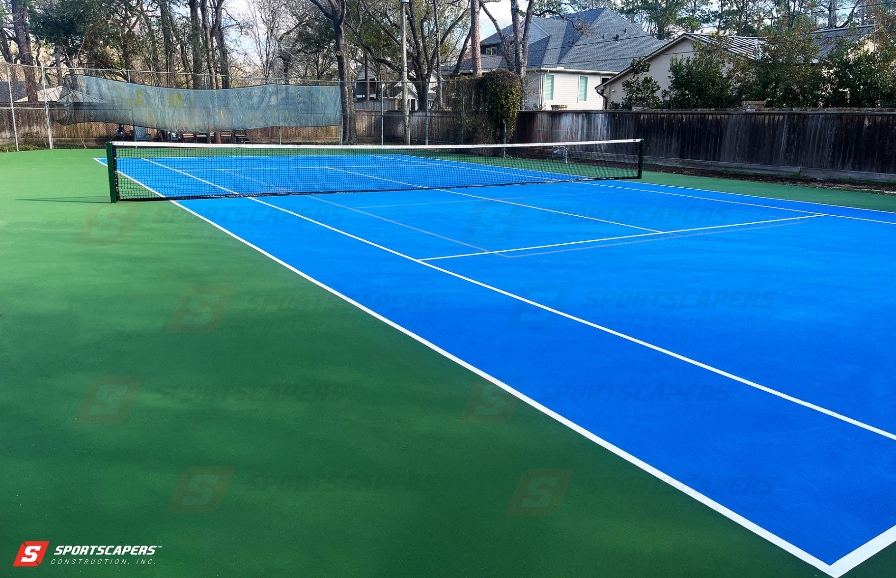 Newly constructed residential tennis court featuring a vibrant blue playing surface with green outer zones in a suburban backyard setting.