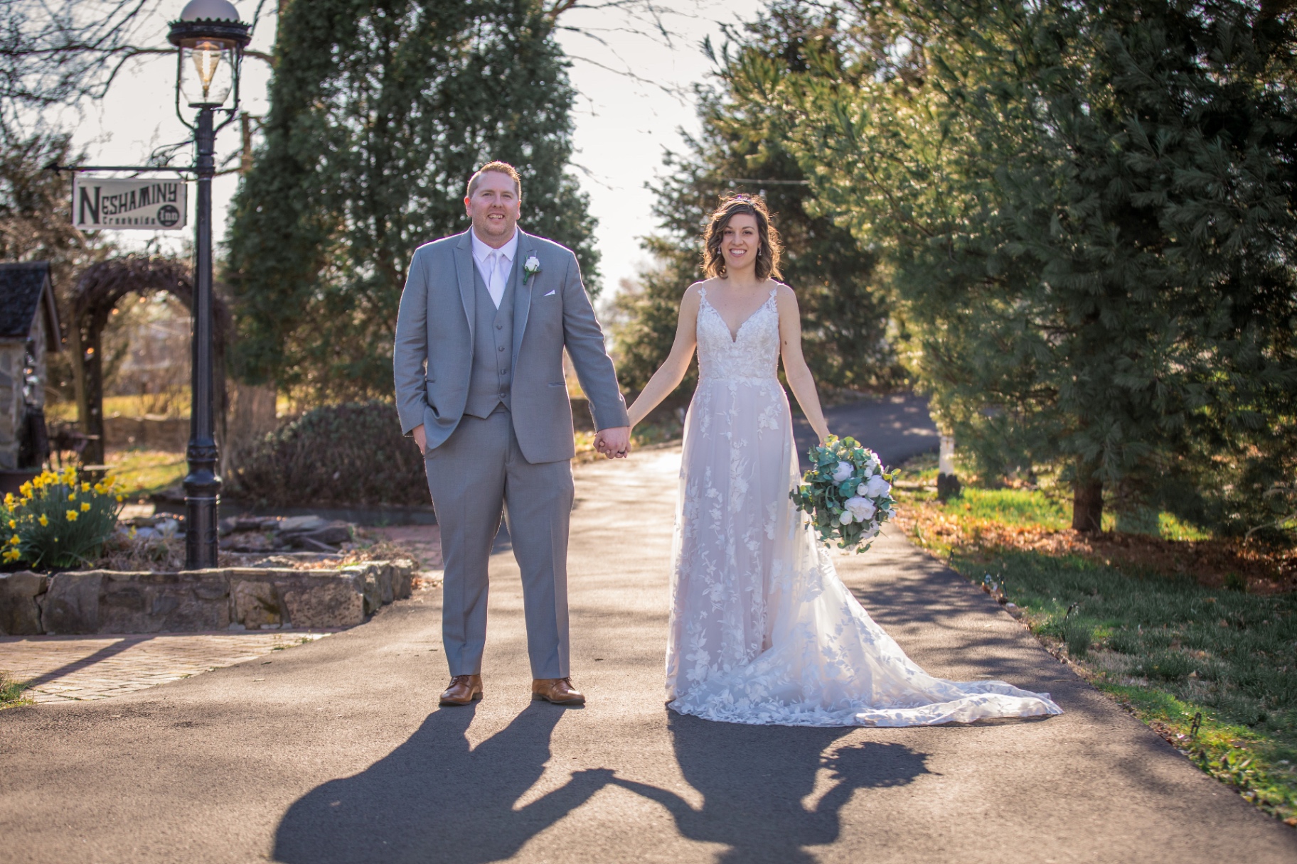 Bride and groom stand hand-in-hand on a sunlit path at Neshaminy Creekside Inn, surrounded by greenery