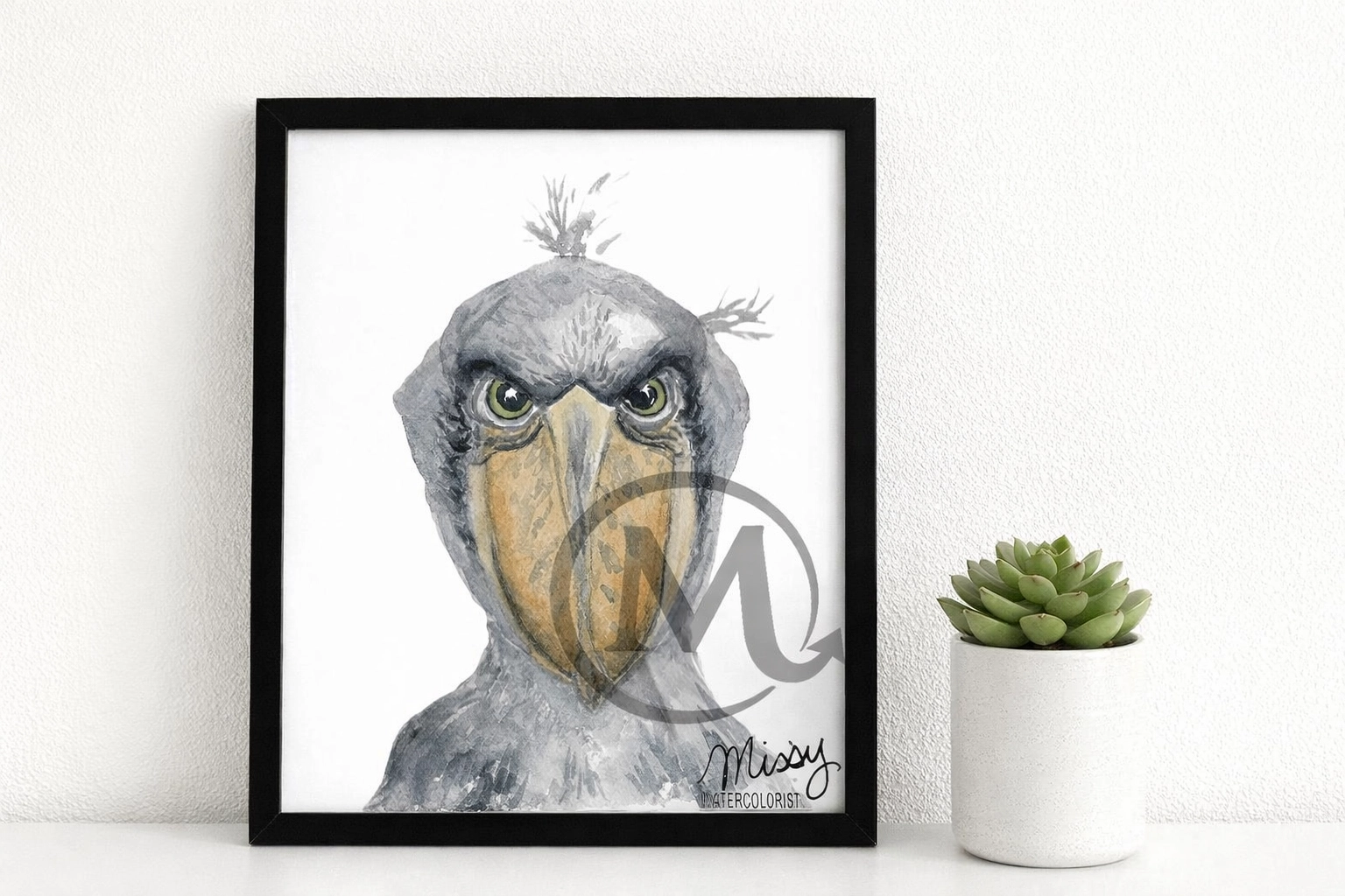 Missy’s original Shoebill watercolor artwork displayed inside a simple black frame mockup on a shelf next to a small potted succulent, with Missy’s watermark and signature fully visible.