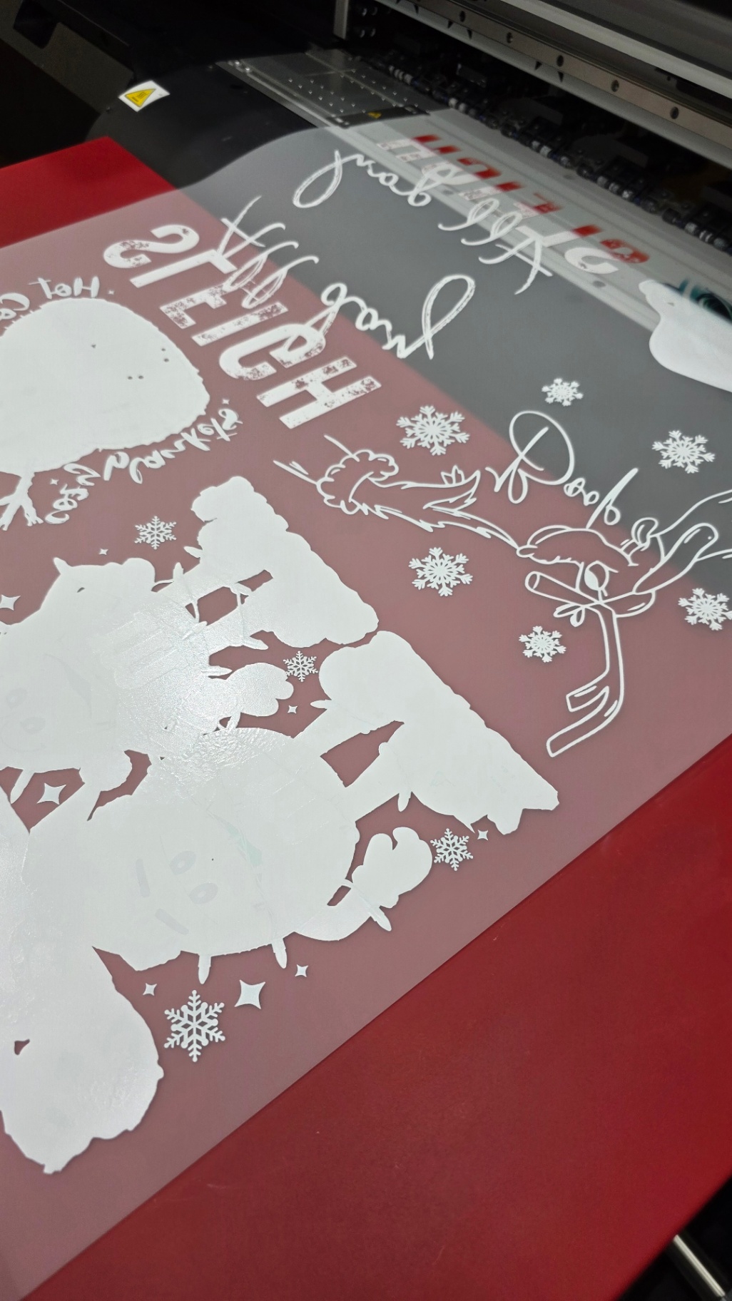 DTF gang sheet printing A DTF gang sheet is printed with various winter and holiday-themed white designs, text, and graphics on a clear film. Positioned on a flatbed printer, it demonstrates Press Ready DTF's ability to deliver clean, detailed, and press-ready custom transfers.