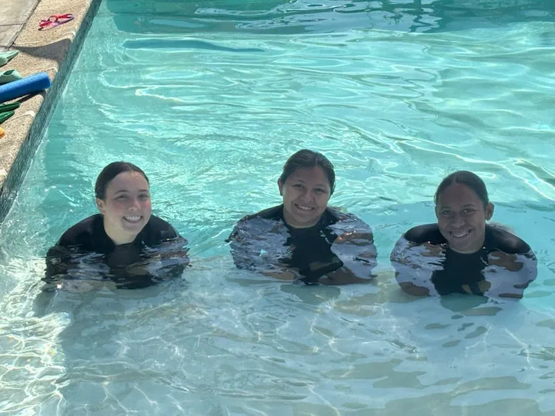 Three professional swim instructors standing in a pool, prepared to support children with diverse needs using adaptive equipment.