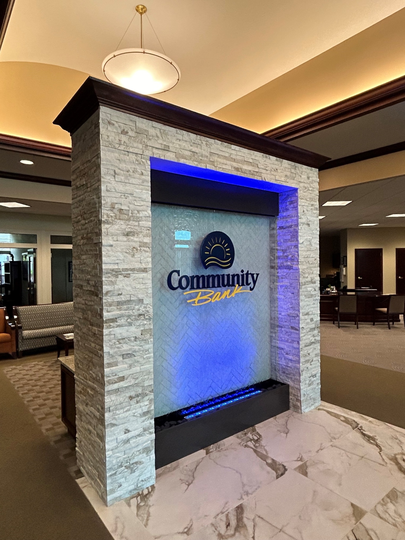Custom-built Indoor Waterwall Installation for Community Bank A modern indoor water feature designed for a bank lobby, featuring a stone wall surround, herringbone-patterned glass, LED accent lighting, and the bank's logo. Utilizes patented, maintenance-free L2O Liquid technology for an impressive and low-maintenance effect that enhances the professional atmosphere and brand image.