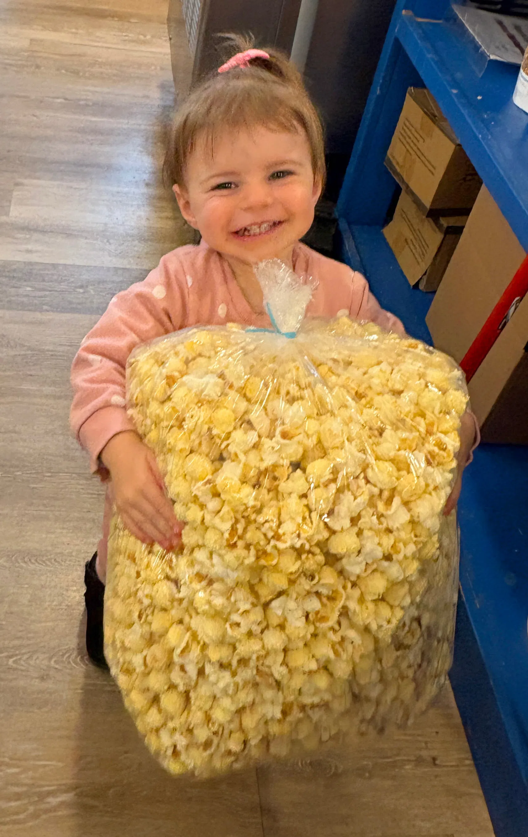 A smiling young child in a pink polka-dot sweater holds a giant clear bag filled with fresh Smith Family Popcorn.