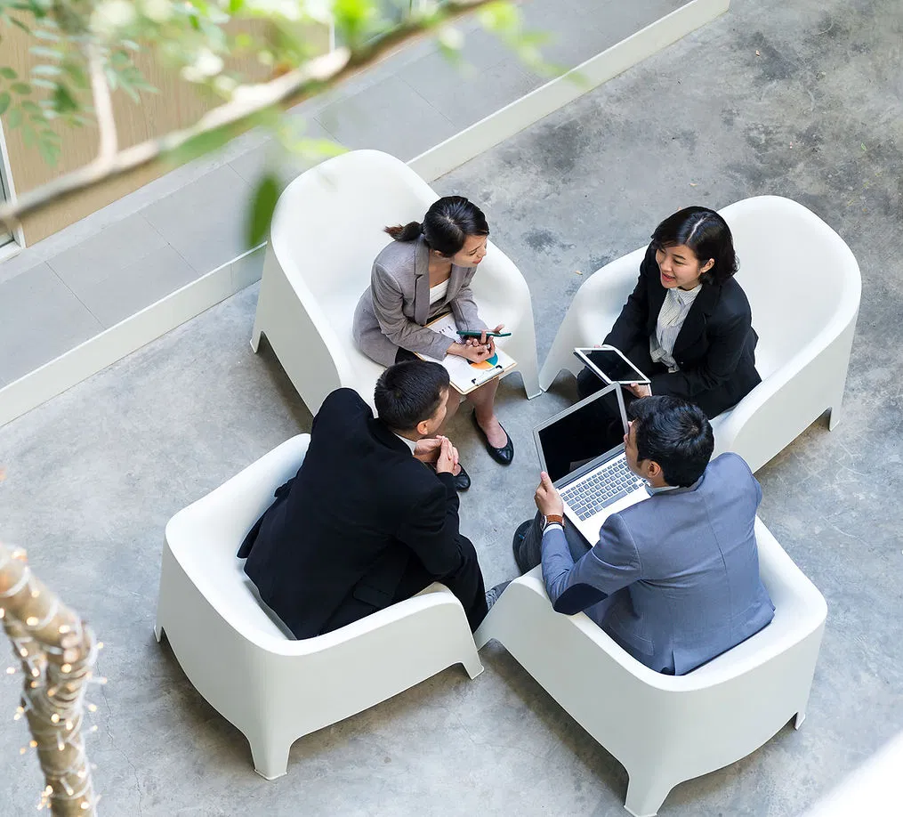 Four business professionals in formal attire are seated in a circle of modern white chairs, engaged in a focused discussion with laptops, tablets, and notebooks, reflecting collaborative problem-solving.