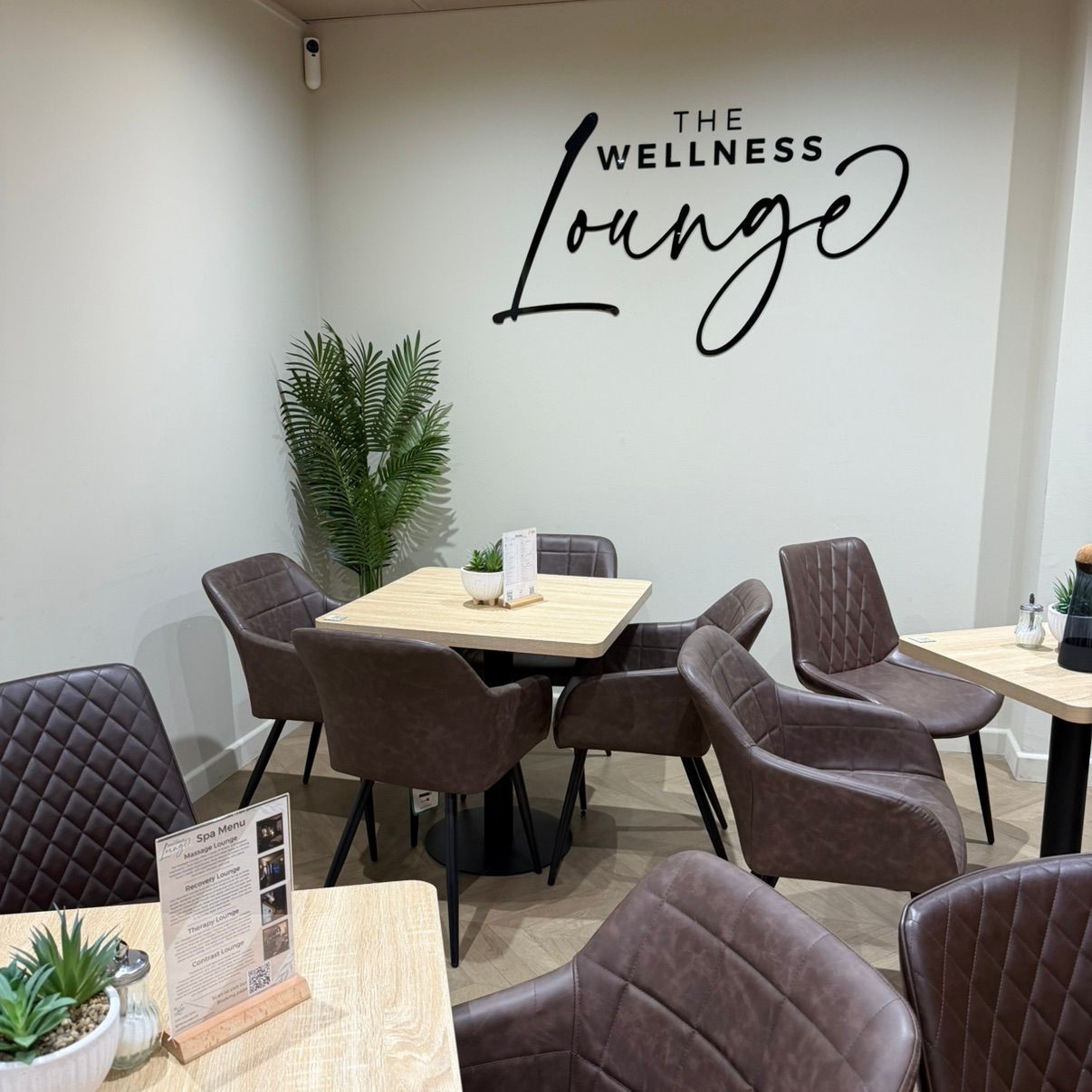 View of the seating area in The Wellness Lounge with modern chairs and tables, featuring the company logo sign on the wall and a succulent on each table.