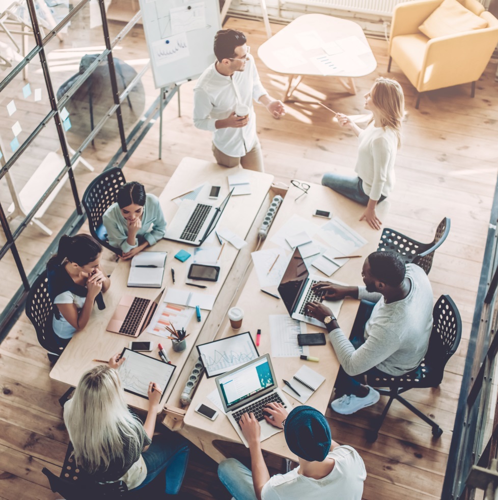 Team Collaboration in Modern Workspace A team collaborates around a large table with laptops, tablets, charts, and documents, actively discussing strategies and analyzing data in a modern, open workspace. Whiteboard and notes indicate ongoing problem-solving, idea sharing, and project planning related to organizational development and employee engagement.