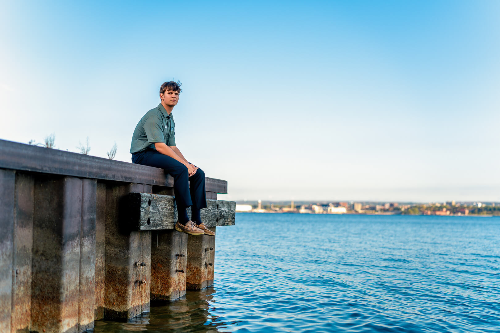 A young man in a green shirt and dark pants sits on the edge of a dock overlooking a calm blue lake, captured in vibrant natural light.
