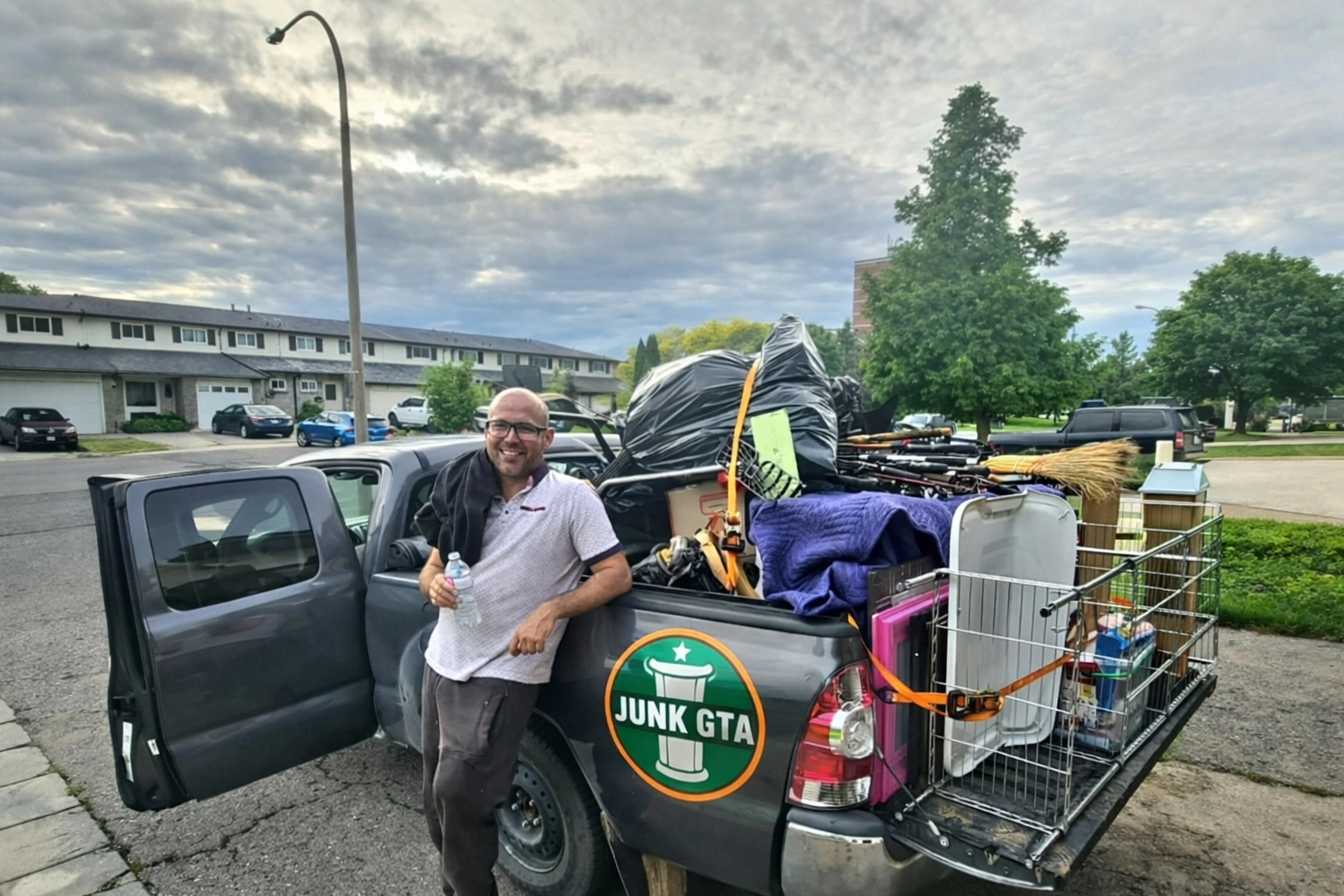 Professional Junk Removal vs DIY Innisfil: The Final Verdict A Junk GTA crew member stands beside a pickup truck loaded with various junk items, such as large bags, a broom, a pet cage, miscellaneous furniture, and household goods.