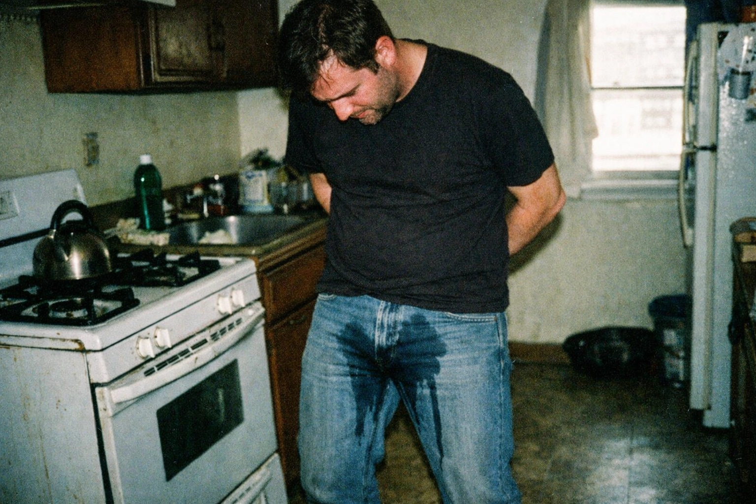 A man in faded denim with a clear wet stain spreading down the jeans — damp texture, loss of control, real-world lighting.