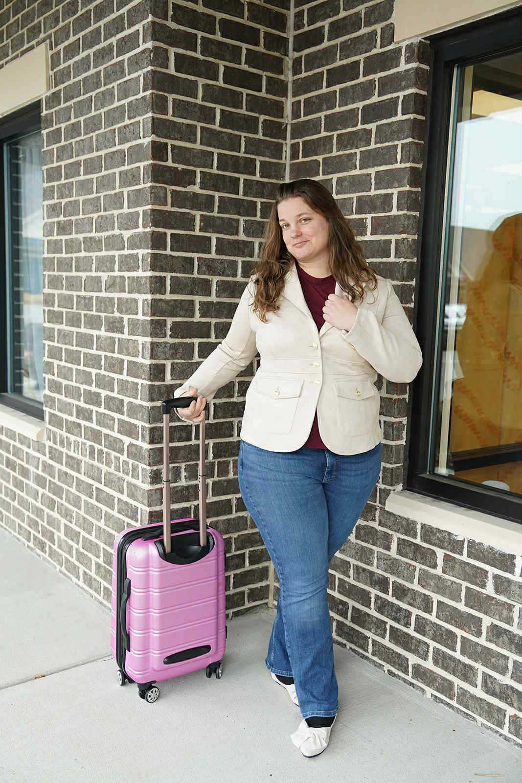 Woman ready for travel outside building A woman dressed in a beige blazer, jeans, and flats stands outside a building with a pink rolling suitcase, prepared for a personalized travel experience arranged by Freedom to Explore Travel Agency.