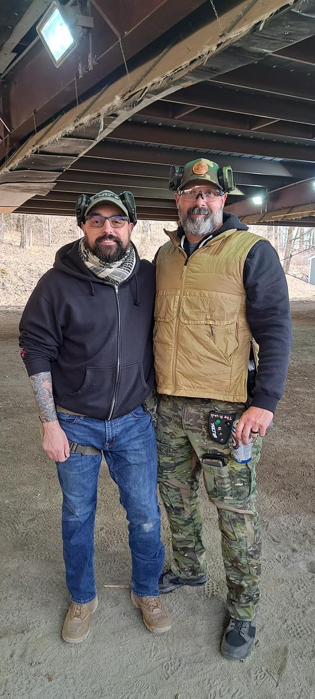 Two adult men at an outdoor shooting range under a metal structure, both wearing hearing protection. One is casually dressed with a hoodie and scarf, while the other wears tactical gear, safety glasses, and camouflage powers.