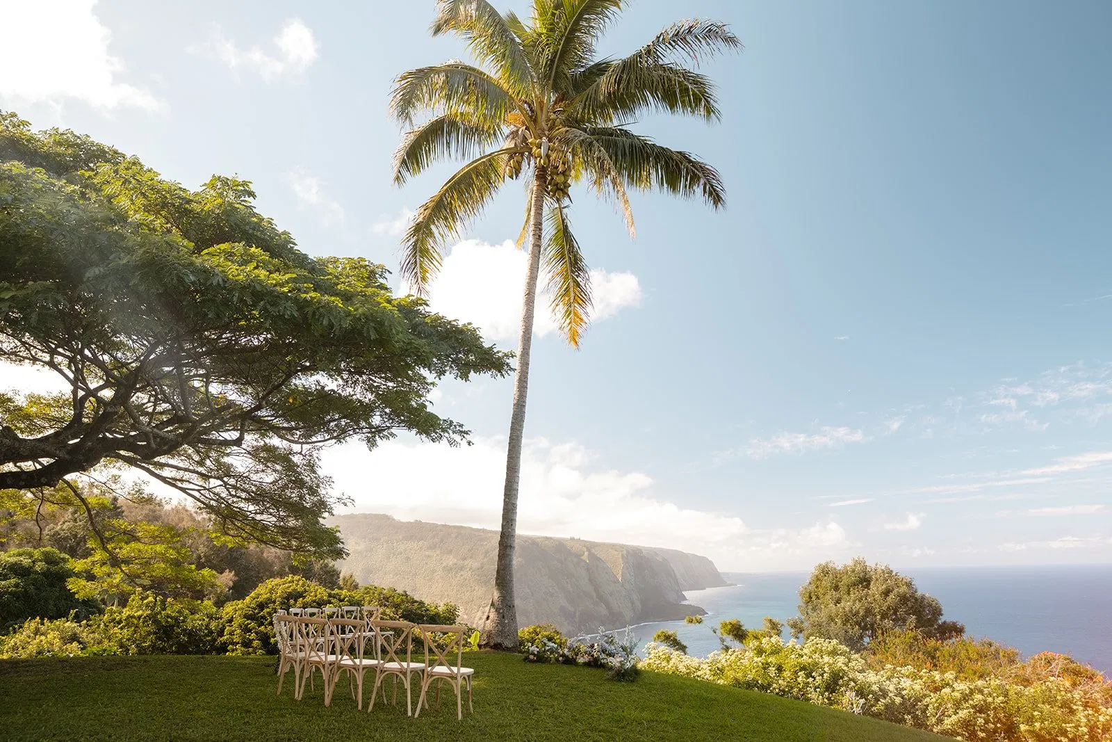 A couple stands on a high, grassy cliff overlooking a dramatic emerald valley and the deep blue Pacific Ocean, with jagged volcanic cliffs in the distance. The lighting is soft and golden, highlighting the organic textures of the grass and the couple's linen attire.