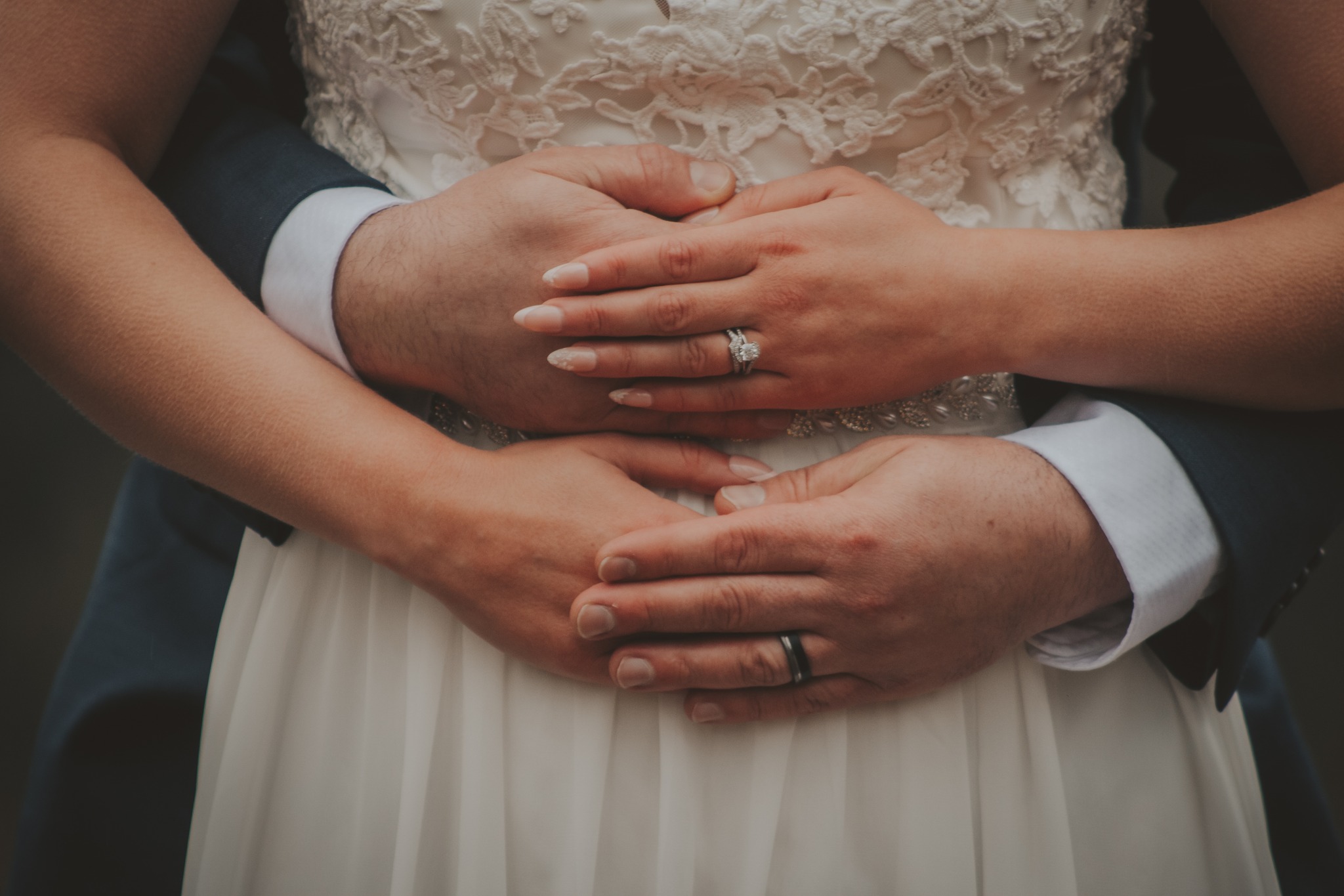 Both are wearing wedding rings, with the detail of lace and suit sleeves visible.