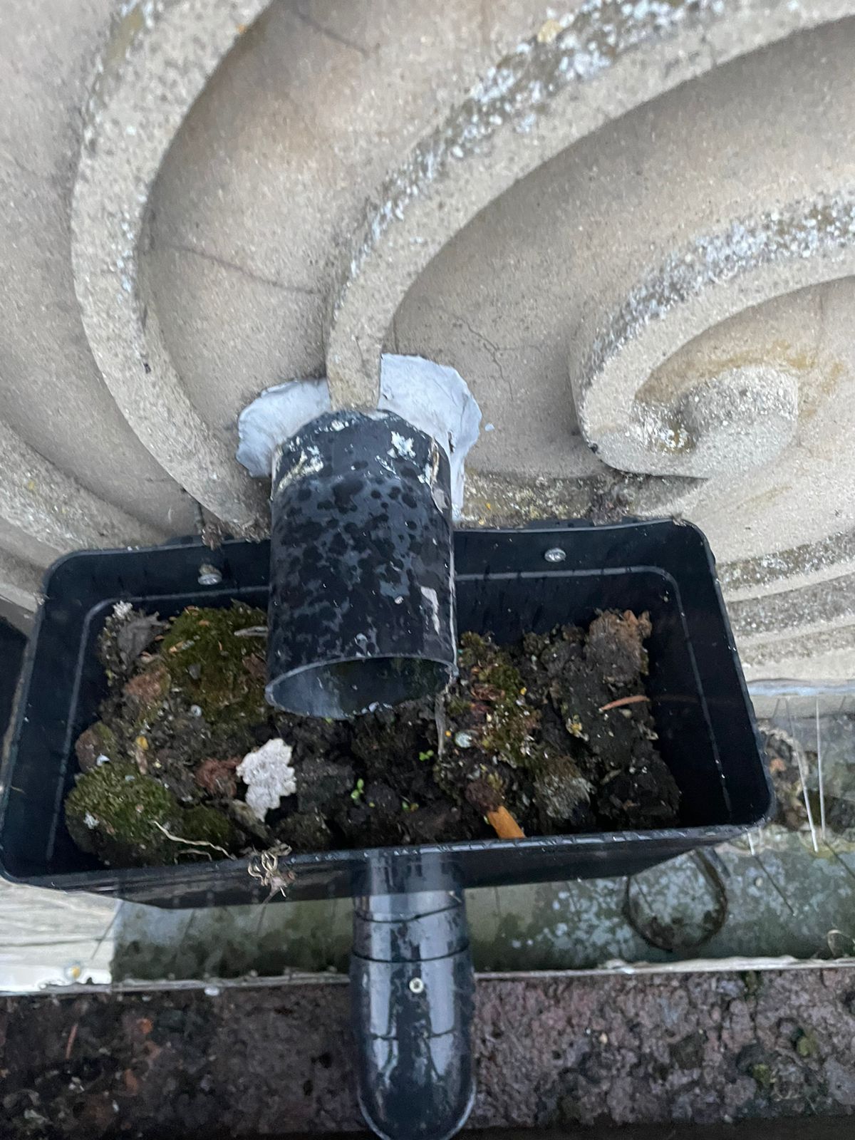 A close-up of a gutter outlet and downpipe heavily blocked with mud, moss, and debris.