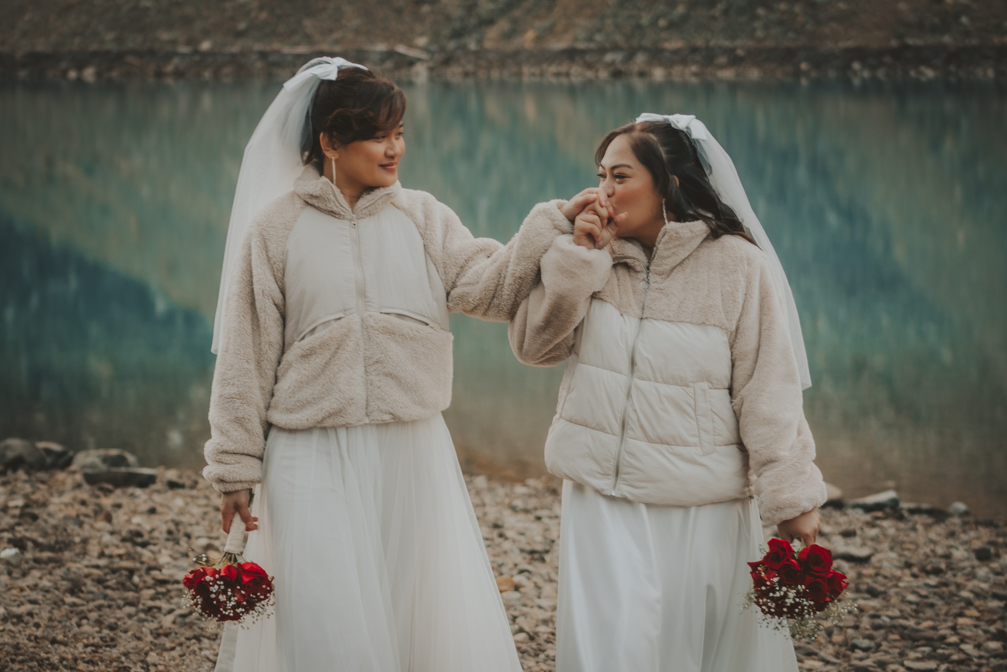 Two brides in matching white gowns and cozy beige jackets at lakeside