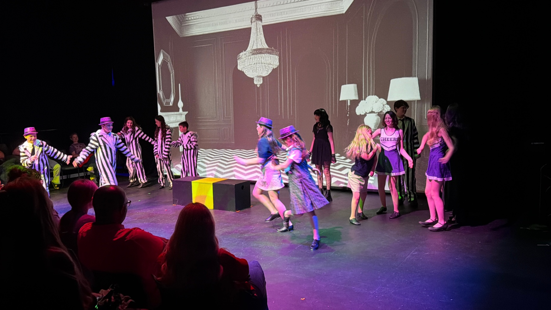 Musical Theater Production Performance Students perform on stage wearing colorful costumes such as black and white striped suits, cheerleader uniforms, and fun hats. The stage features a projected set design of an elegant room in the background while audience members watch a lively group scene.