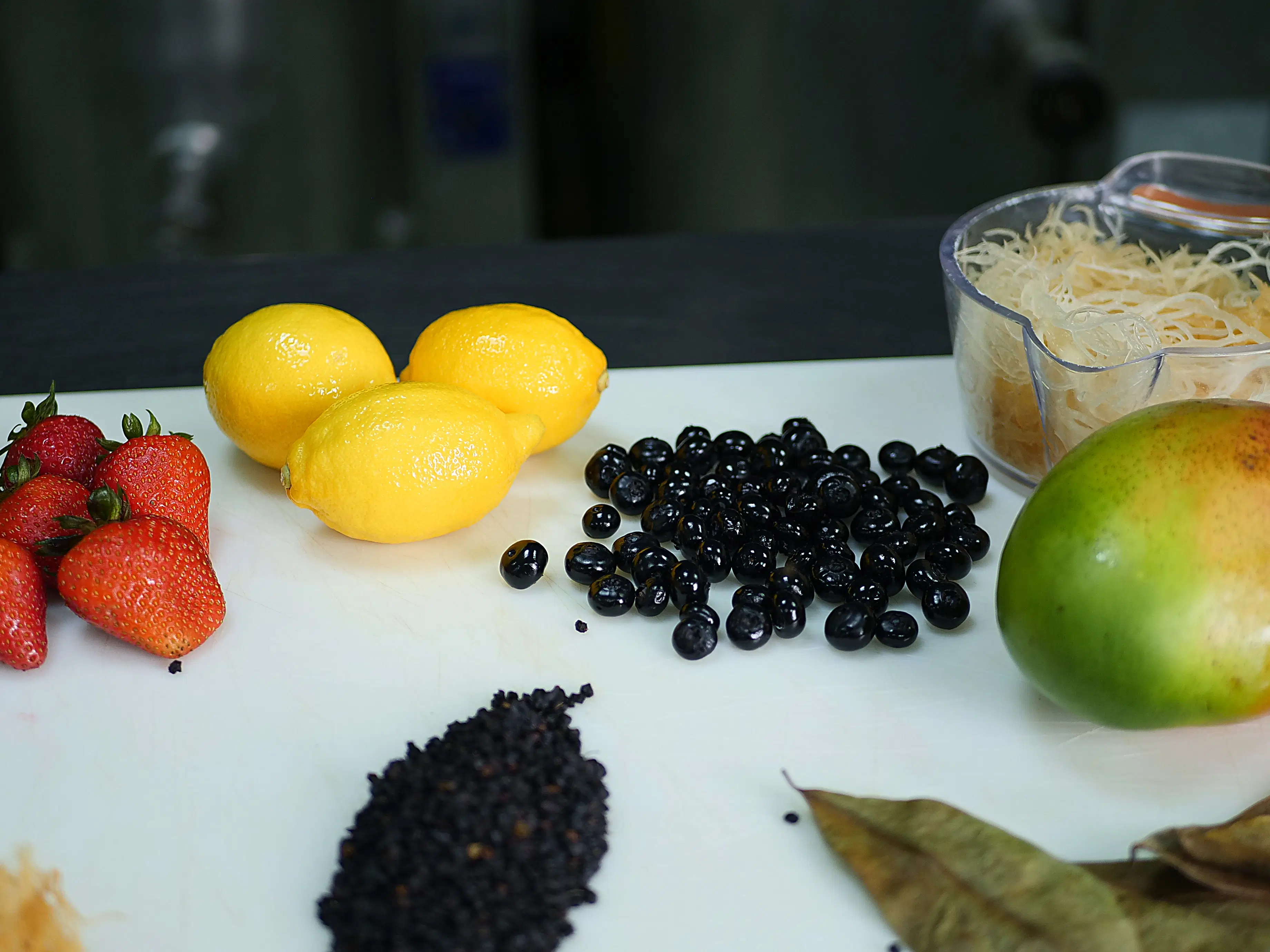 A vibrant assortment of fresh ingredients used for Sea Moss Gel flavors, including strawberries, lemons, blueberries, elderberries, mango, raw gold sea moss, and dried leaves, artfully arranged on a preparation surface to showcase the premium, natural components in Ms. D’s Wellness Goods products.