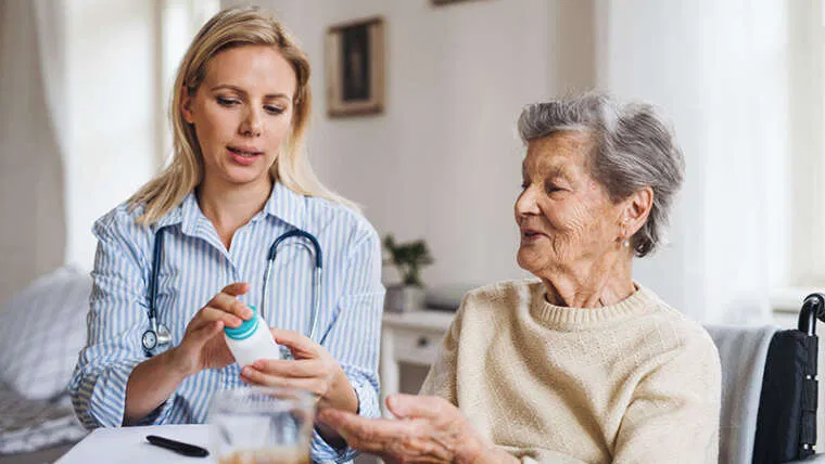 A Registered Nurse explains and dispenses medication to a senior woman in a home setting, demonstrating compassionate one-on-one care and personalized medication management