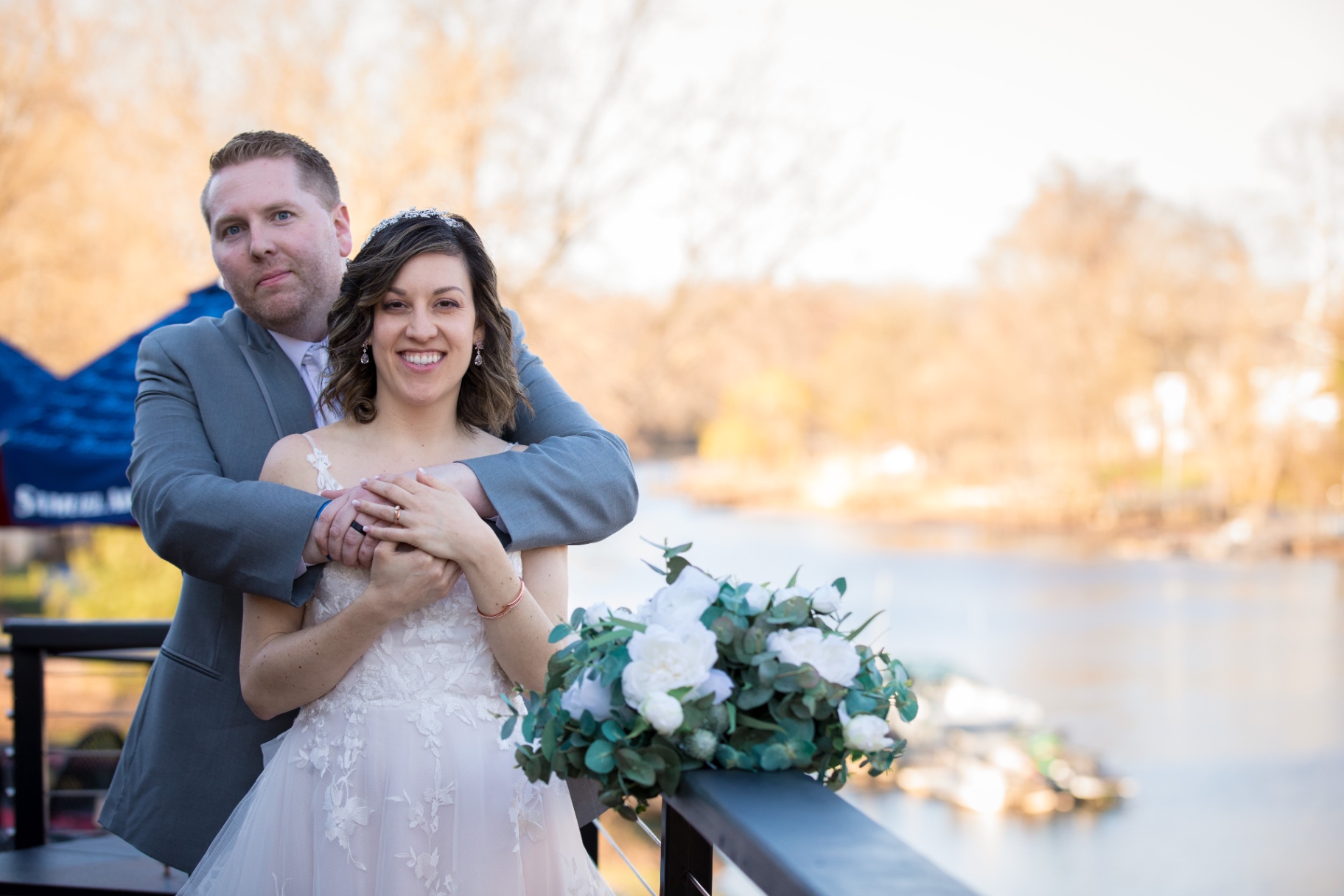 A bride and groom embrace and smile on an outdoor balcony overlooking the water, bouquet in the foreground