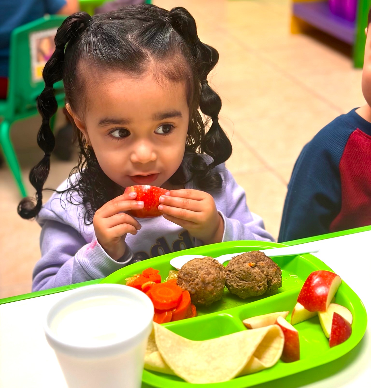 A young child sits at a table eating meatballs, sliced apples, carrots, and a tortilla, with a cup of milk, in a brightly lit classroom. The scene reflects an emphasis on nutrition and wellbeing as part of early childhood learning programs.