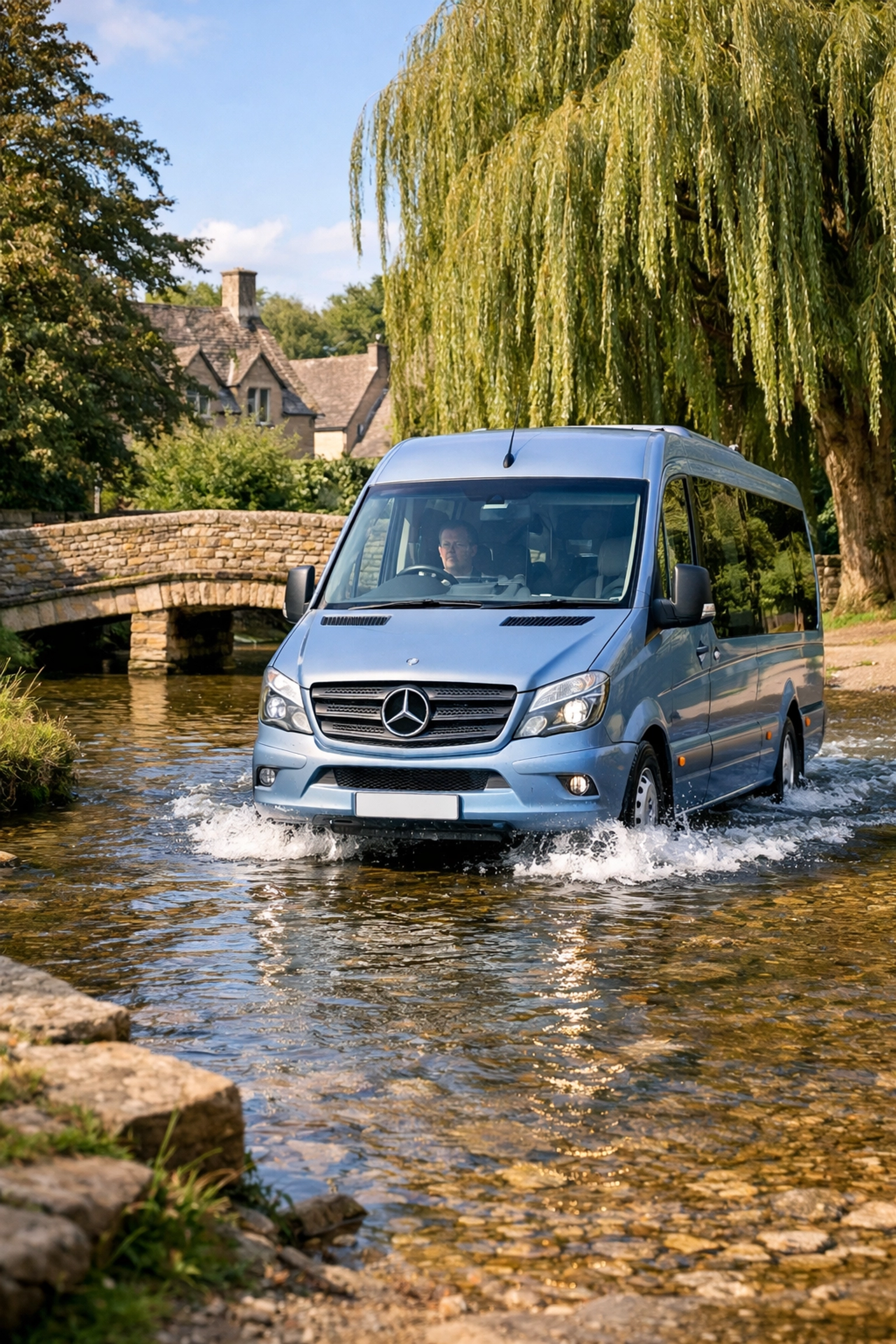 A silver blue Shakespeare Coaches minibus driving through the famous river ford in Upper Slaughter.