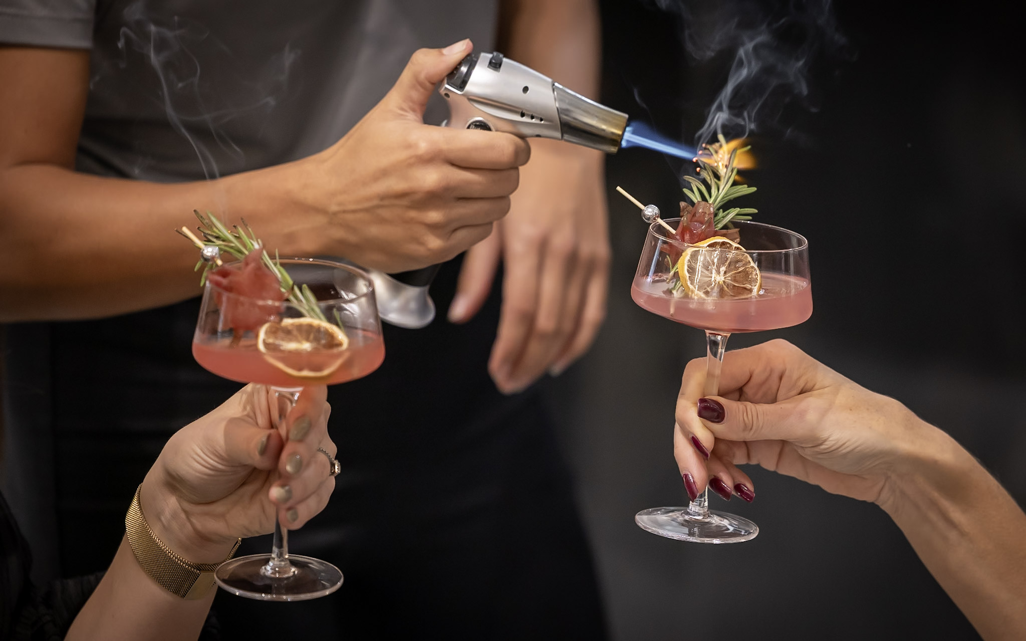 A bartender uses a culinary torch to flame garnish on two cocktails