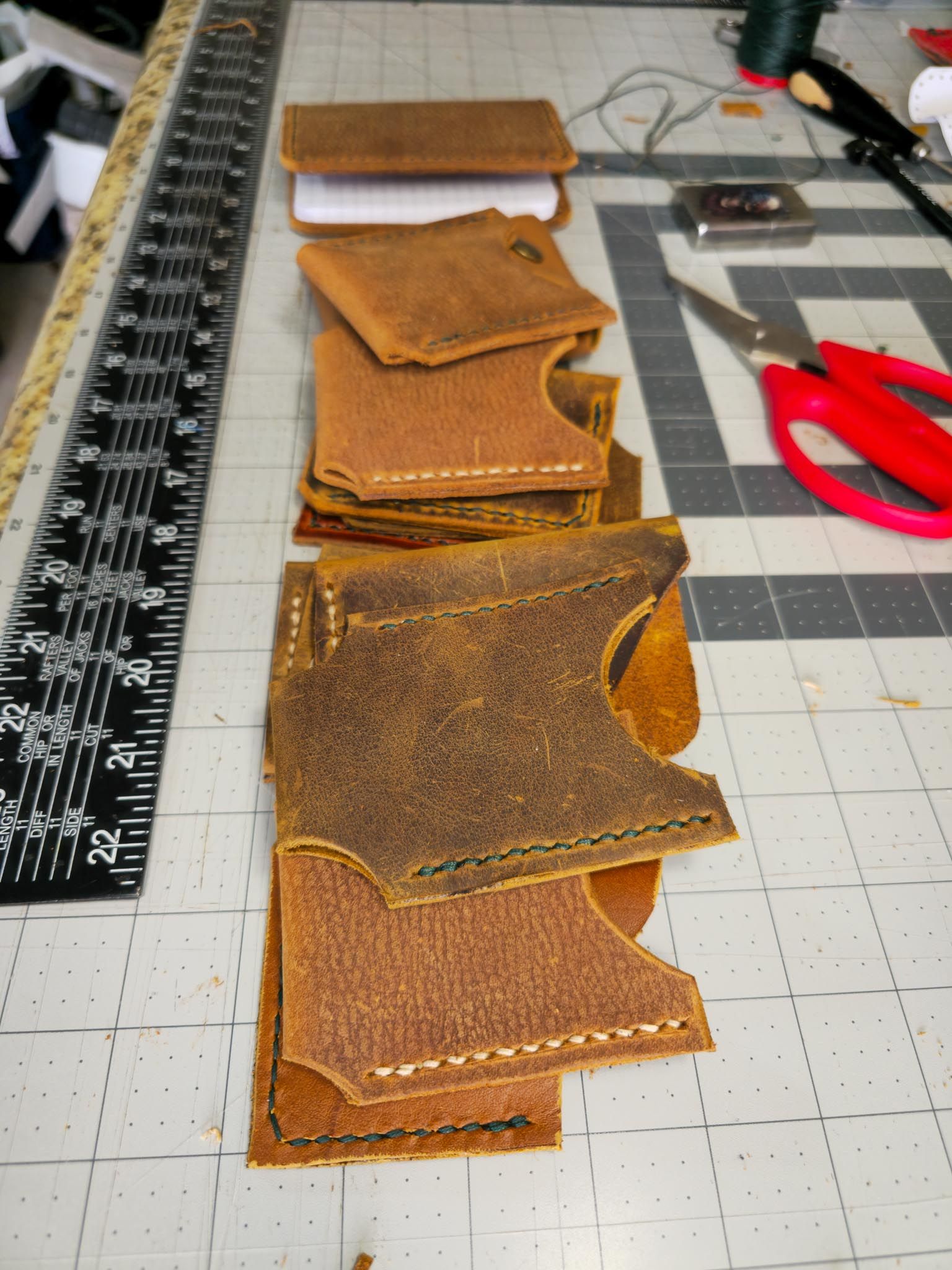 A row of rugged brown leather card sleeves with visible hand-stitched details lined up on a workbench. Leathercraft tools, a ruler, and thread are scattered around.