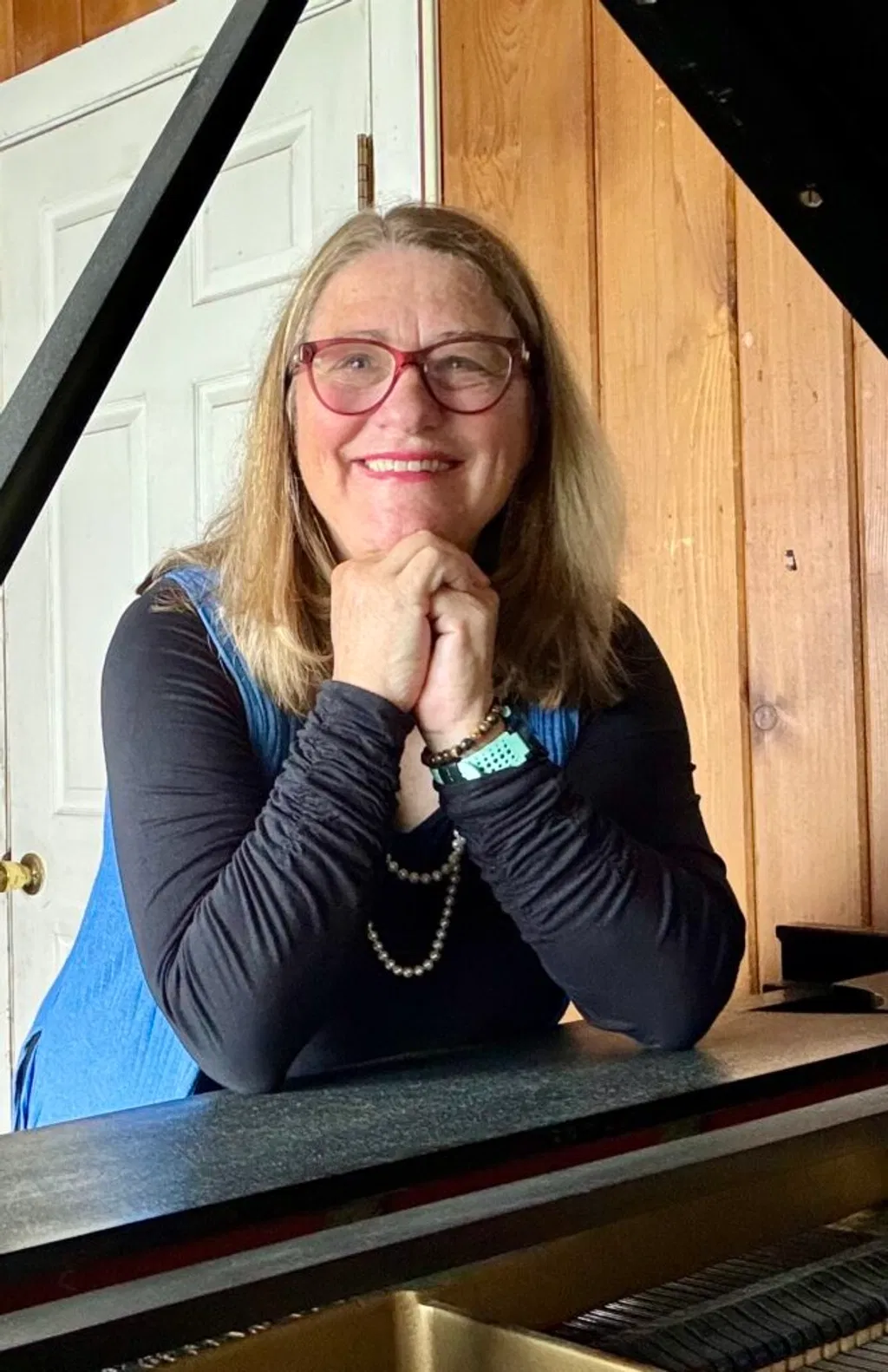 Smiling woman with glasses and long hair, wearing a blue vest, black top, and a pearl necklace, leaning on a grand piano with hands folded under her chin.