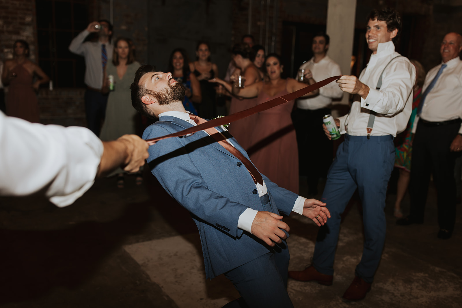 Wedding guests enjoy a lively dance floor moment as one person in a suit participates in a limbo game, surrounded by smiling friends with drinks, capturing the energetic party atmosphere created by JAMMIN' DJs' entertainment services.