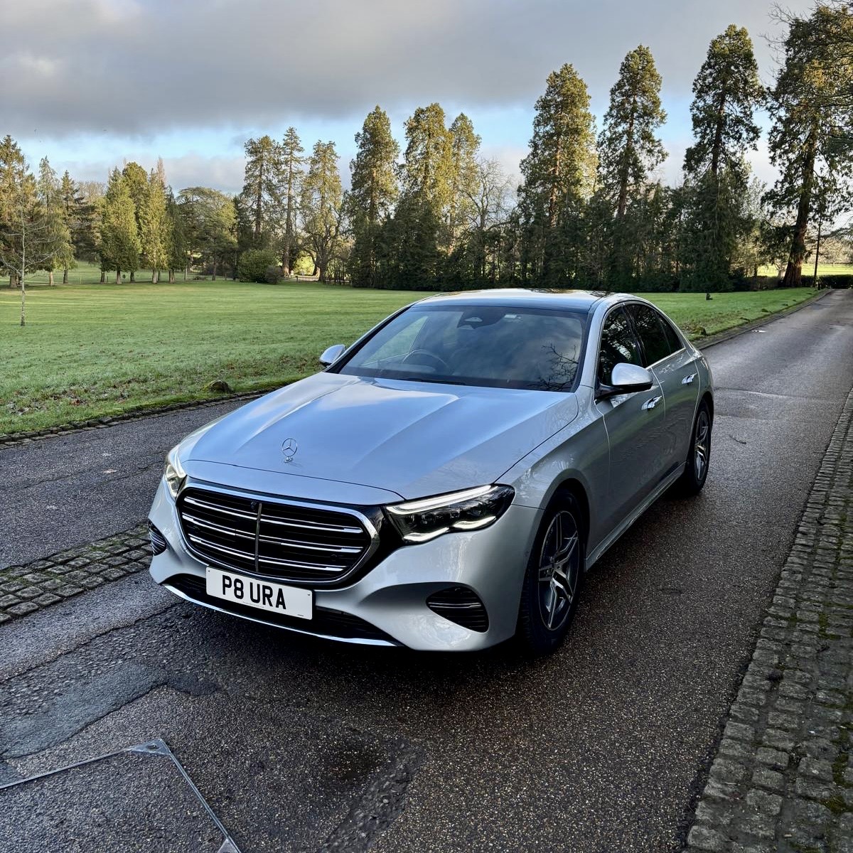 A silver Mercedes-Benz executive saloon parked on a quiet, tree-lined driveway, representing Aura Journeys’ luxury chauffeur fleet.