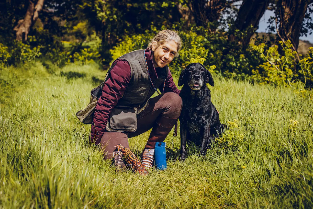 A gundog trainer kneels beside a black Labrador in a grassy field, demonstrating 1 to 1 gundog training.