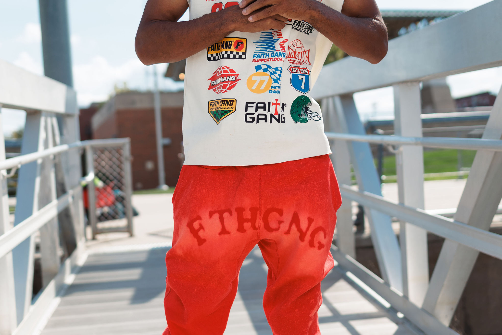 TruVine Apparel Faith Gang Streetwear Outfit Close-up of a model wearing TruVine Apparel's red acid wash joggers with bold "FTHGNG" (Faith Gang) text across the front, paired with an off-white t-shirt featuring multiple colorful Faith Gang racing-inspired graphics and logos, showcasing the brand's streetwear Christian message and premium design details.