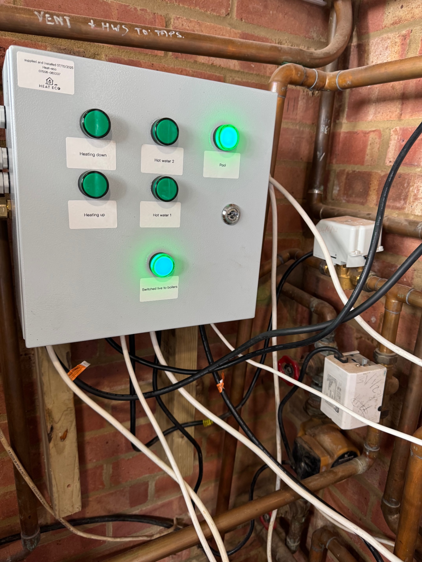 Wall-mounted control panel installation by Heat Eco Building Services Managing heating, hot water, and pool systems. Unit features labelled push-button controls, active indicator lights, copper piping, and neatly routed wiring, demonstrating integration of energy-efficient solutions for residential or commercial settings.