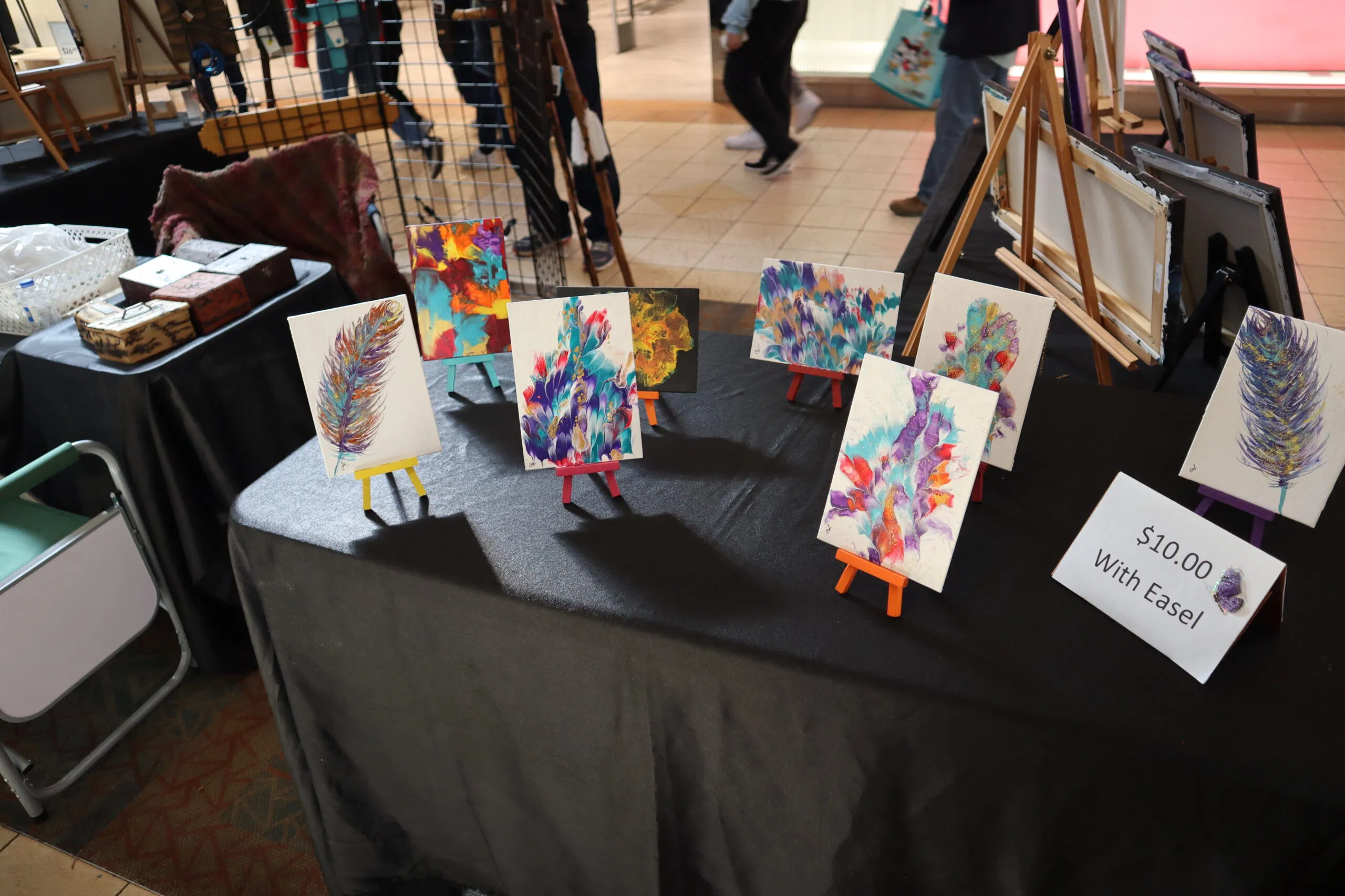Tabletop vendor setup at the ACC Bazaar featuring vibrant, hand-painted feather-themed mini canvases on colorful easels