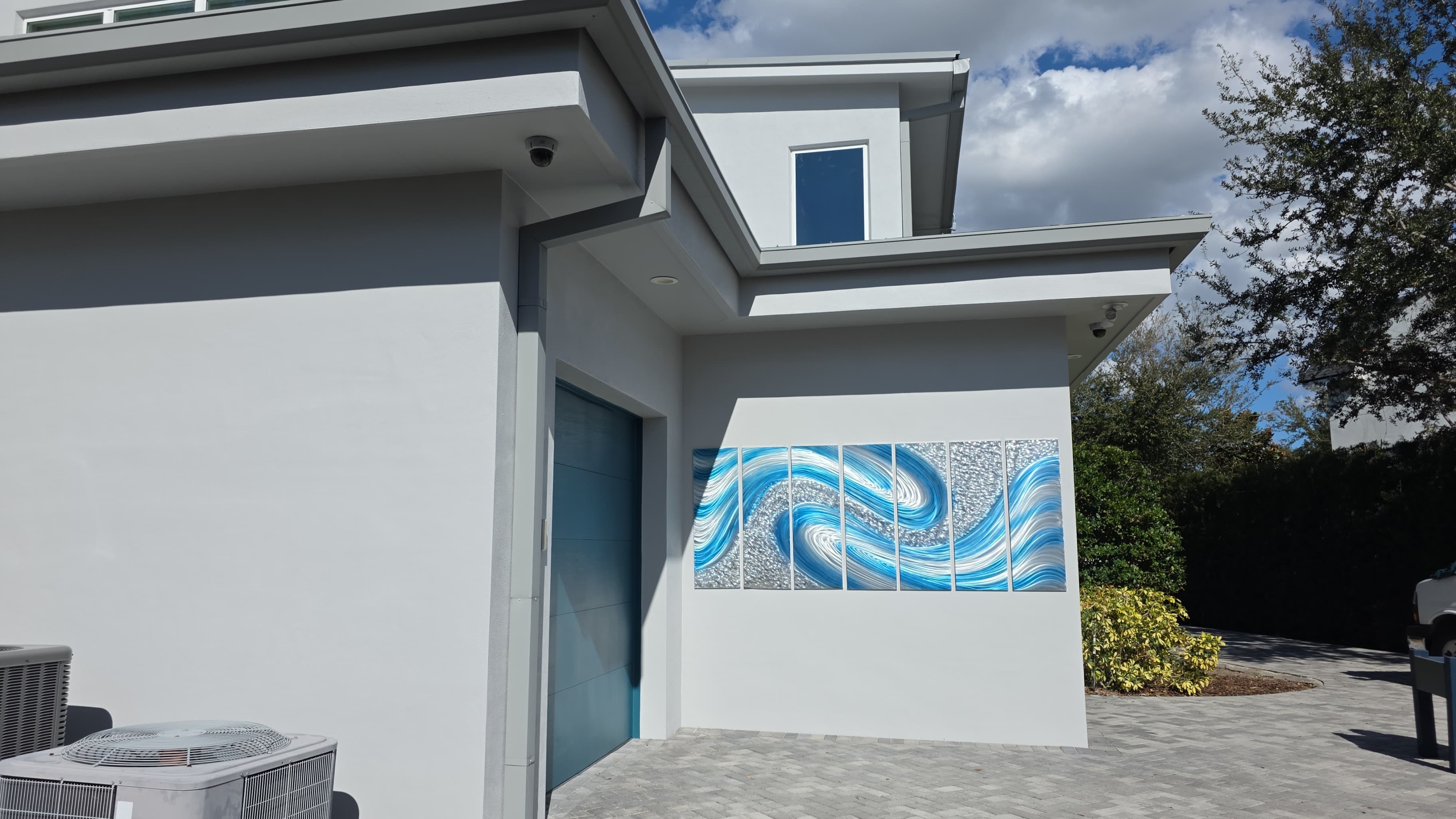 Modern Exterior Home Renovation Modern exterior home renovation featuring smooth, light grey stucco walls, a contemporary blue garage door, and a decorative blue and silver wave-themed mural panel. The driveway is finished with interlocking pavers, and the landscaping includes manicured bushes and trees, highlighting the clean aesthetic and precise craftsmanship.