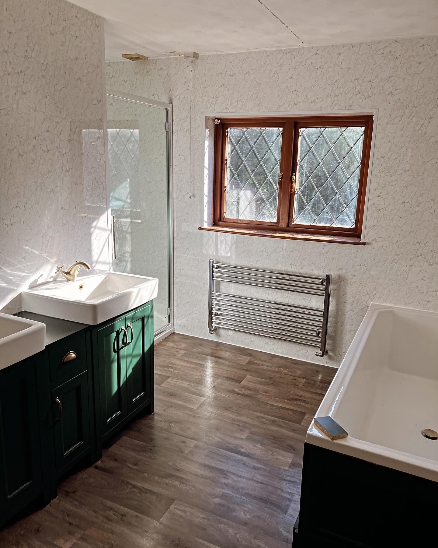 Modern bathroom installation featuring eco-friendly fixtures, dual sinks with stylish green cabinetry, a walk-in shower, energy-efficient chrome towel radiator, wood-effect flooring, and a standard bathtub. All plumbing and heating work completed to high standards for optimal efficiency and comfort.