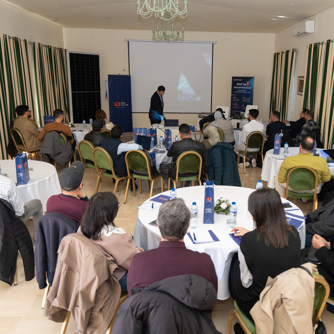 VLD SOLAR Seminar A professional seminar hosted by VLD SOLAR, where participants are seated at round tables watching a presentation about photovoltaic technology. The room features branded materials such as bags and pamphlets on the tables. A company representative presents at the front near a projector screen and banners displaying VLD SOLAR and partner brands.