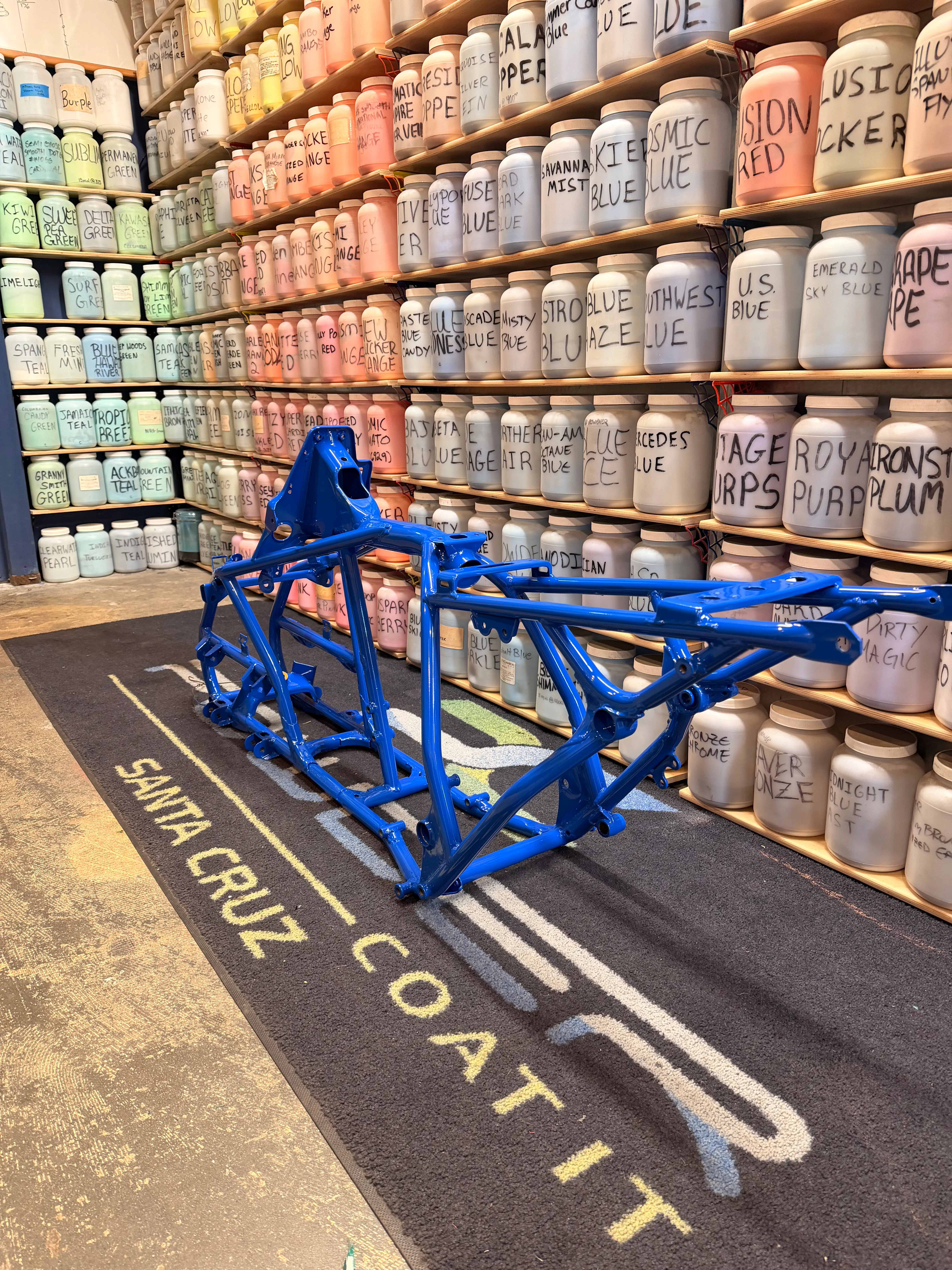 A freshly powder coated metallic blue motorcycle quad frame is displayed on a Santa Cruz Powder Coat It mat, surrounded by shelves filled with labeled jars of powder coating pigments in a wide array of colors, showcasing an extensive color selection and expertise in custom automotive coatings.