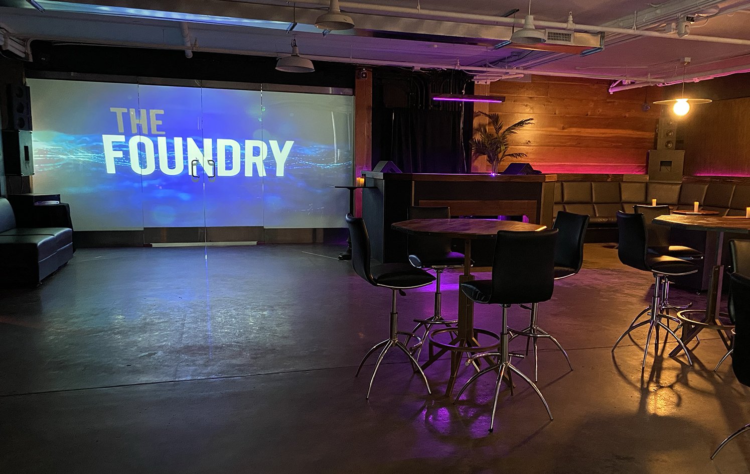 The Foundry Industrial chic event space featuring polished concrete floors, exposed beams, modular round tables, modern bar stools, built-in stage, wraparound booth seating, and customizable lighting