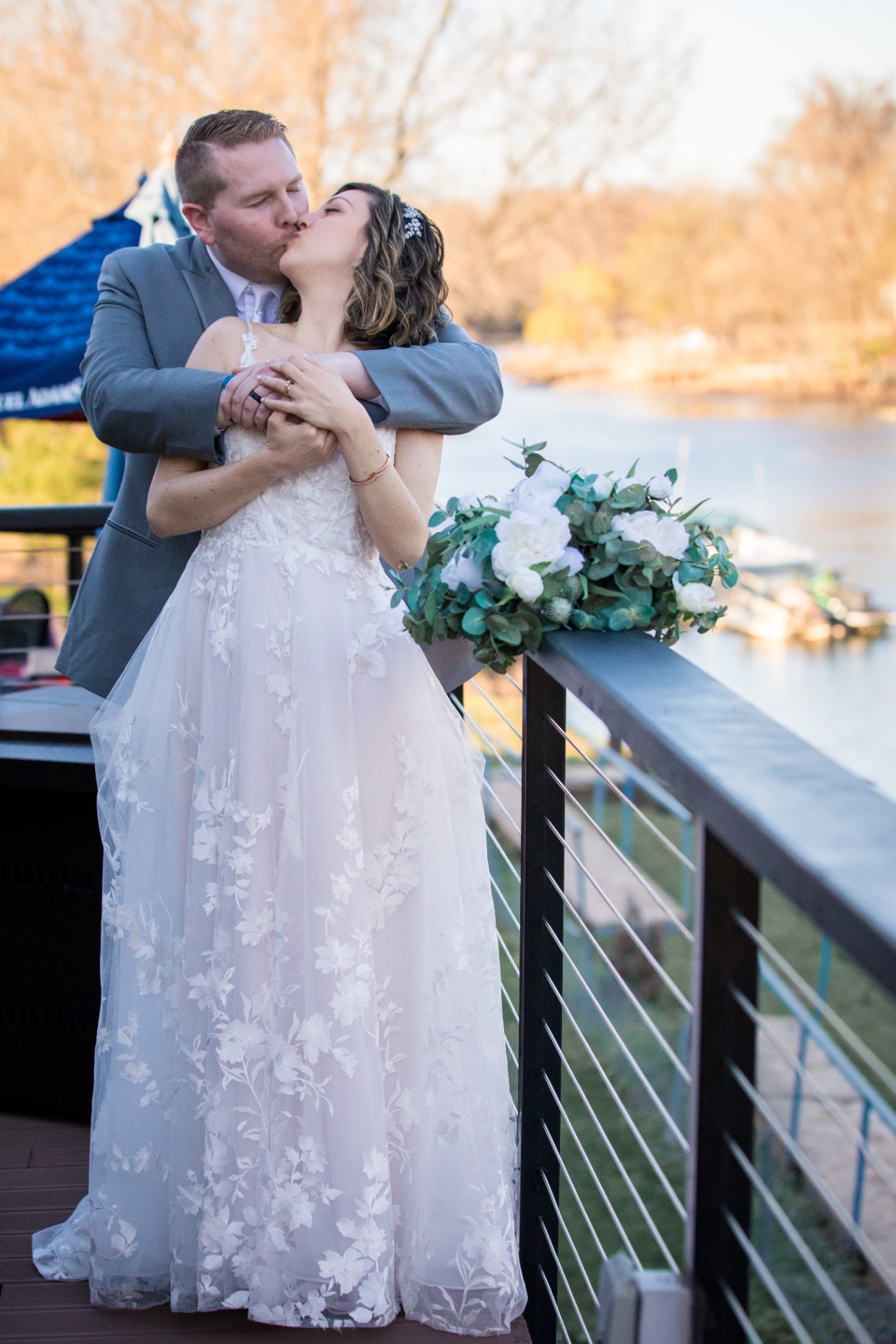 Bride and groom share a romantic embrace and kiss on an outdoor deck, bouquet resting on the railing, river in the background
